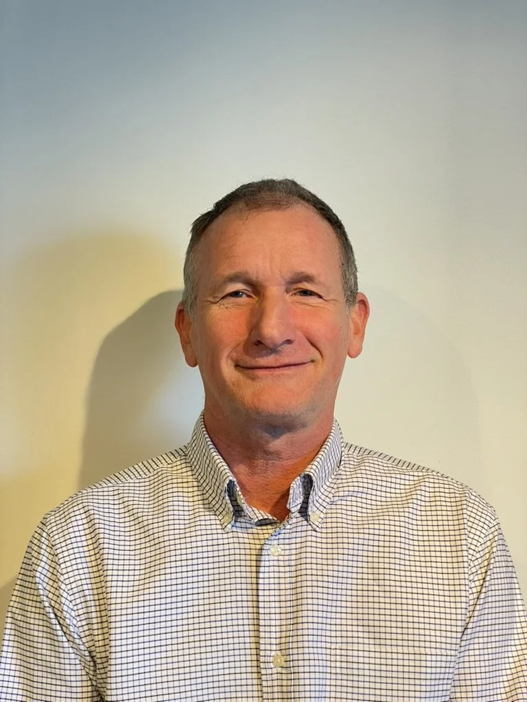 Douglas Moore dressed in bright orange outdoor gear with black sunglasses, a black knit cap, and gloves, holding a rifle outdoors near a leafless tree.