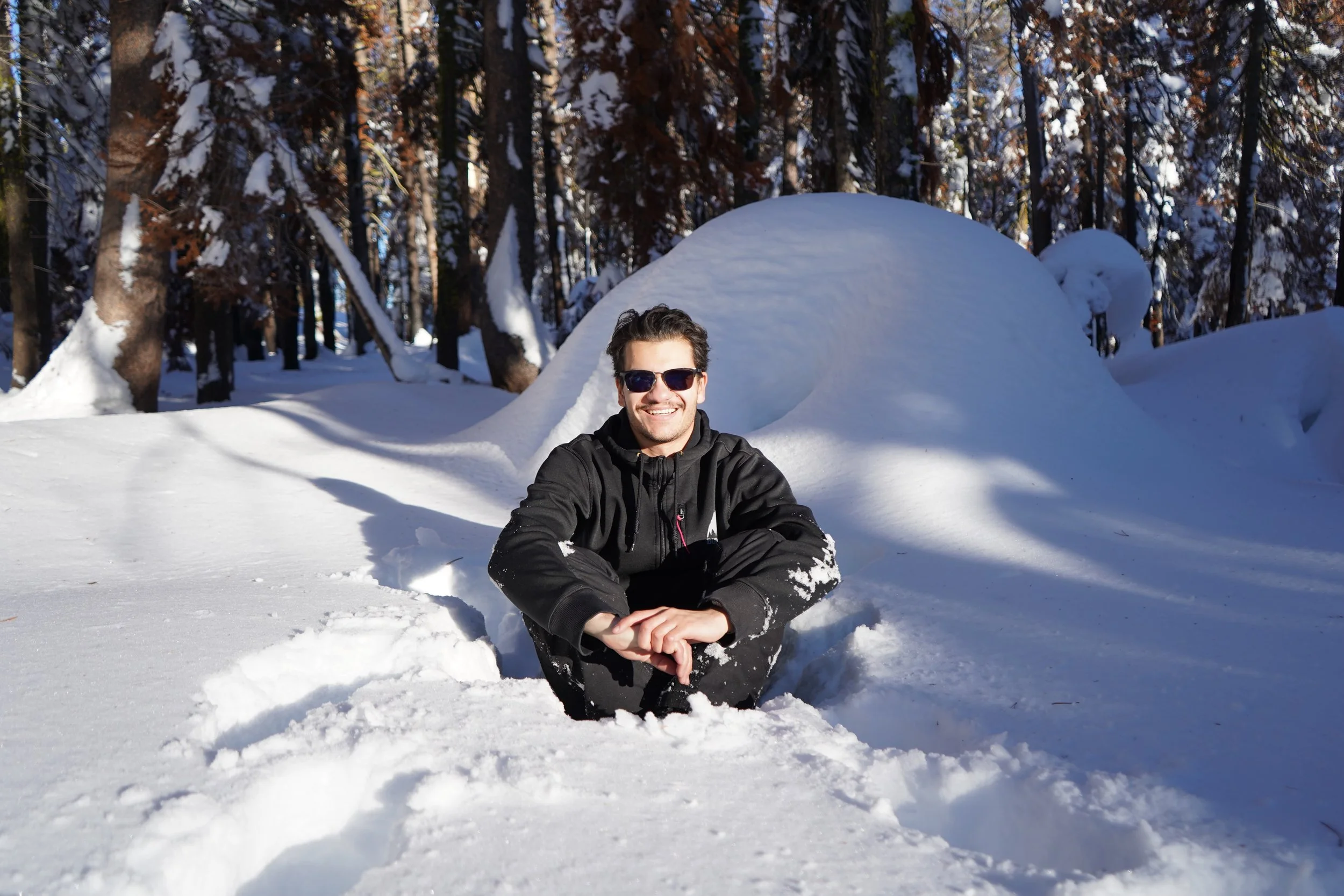 Coby Cruz sitting in the snow in a forest, smiling and wearing dark sunglasses and a black jacket.