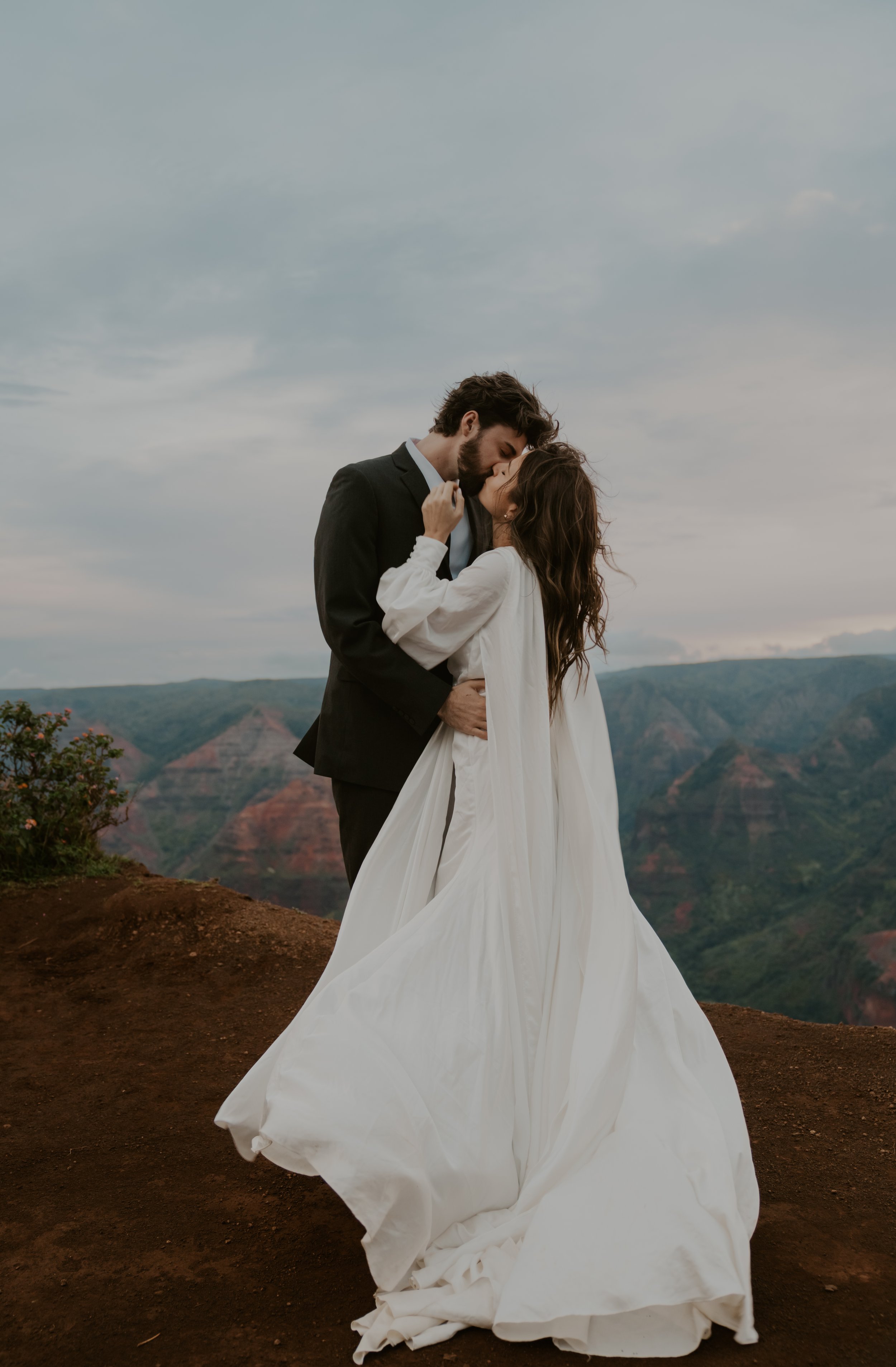 A bride and groom share a kiss on a mountain overlooking a valley with colorful cliffs, the groom in a black suit, the bride in a white wedding dress.