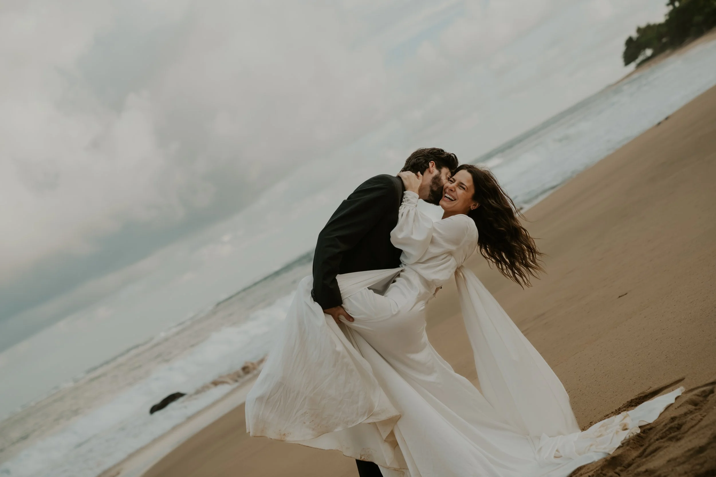 A joyful couple dressed in wedding attire sharing a dance on a beach during cloudy weather.