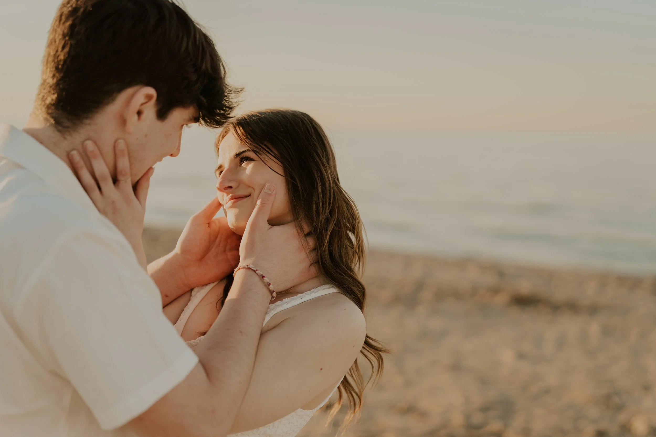 A man and woman on a beach during sunset, holding each other's faces and smiling lovingly at each other.
