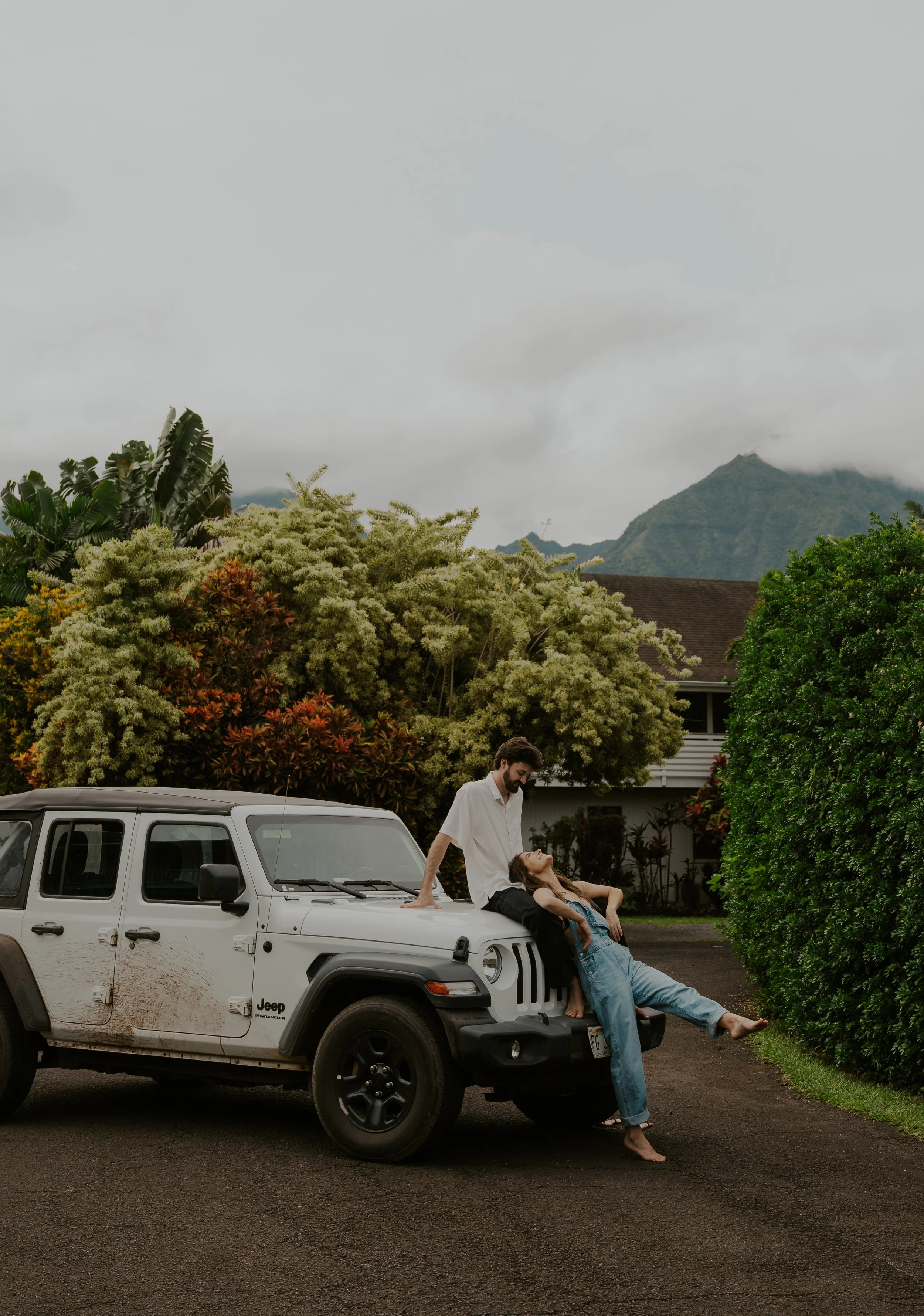 A couple relaxing on the front of a white Jeep with a dirt stain, parked on a driveway in a lush, green area with trees and mountains in the background. The woman is lying against the Jeep with her eyes closed, while the man is standing and leaning o
