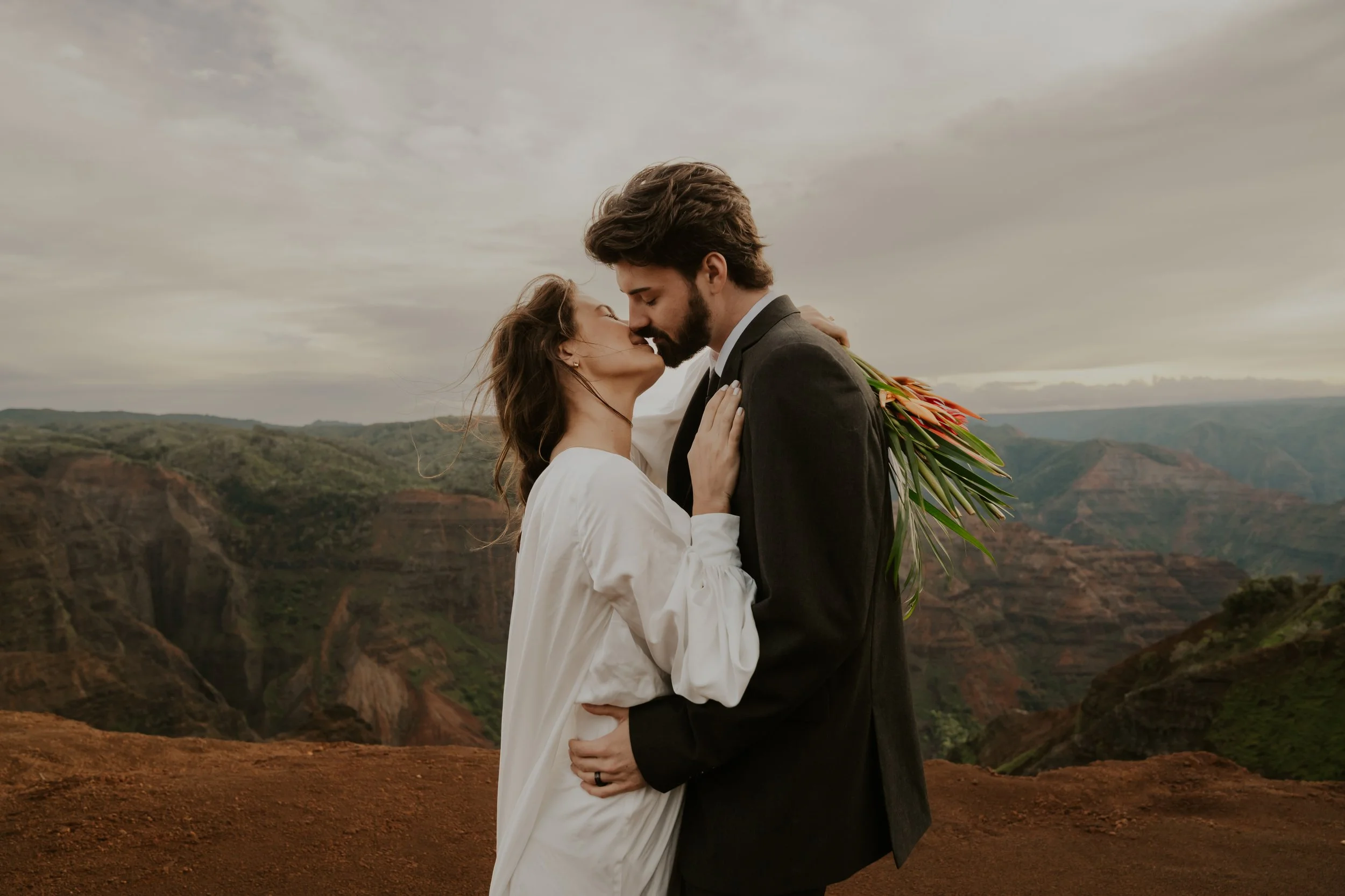 A romantic couple kissing on a mountain overlook, with a woman in a white dress and a man in a black suit, holding a bouquet of flowers, and the background showing a canyon landscape under a cloudy sky.