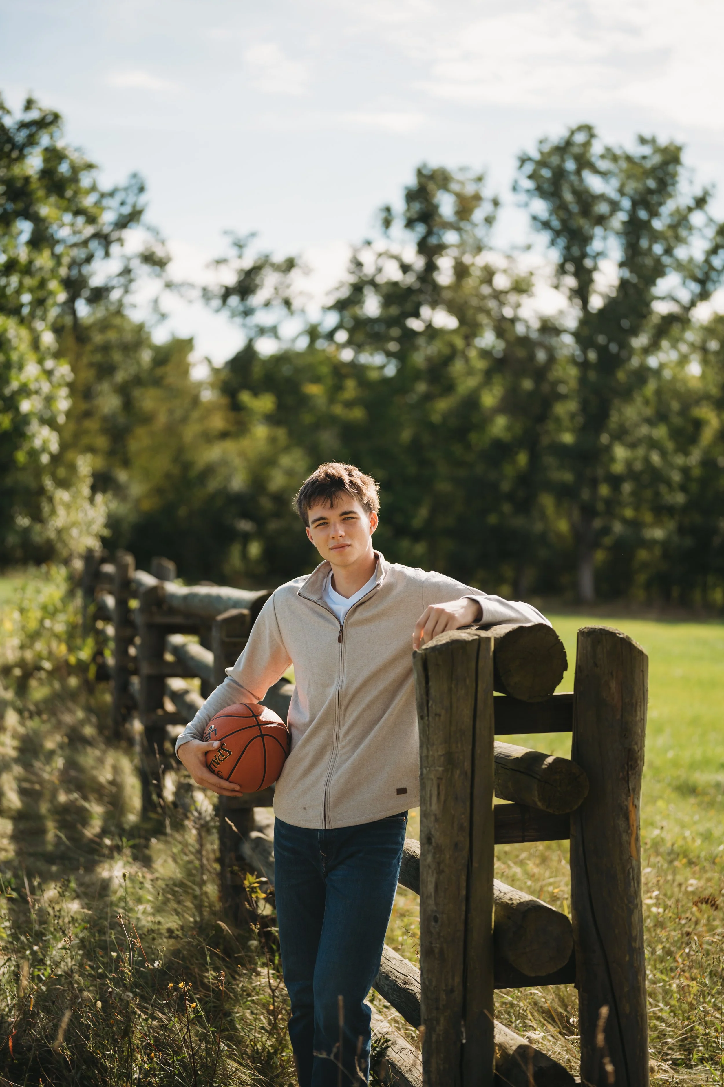 A young man with dark hair holding a basketball stands outdoors by a wooden fence on a sunny day with a background of green trees and grass.