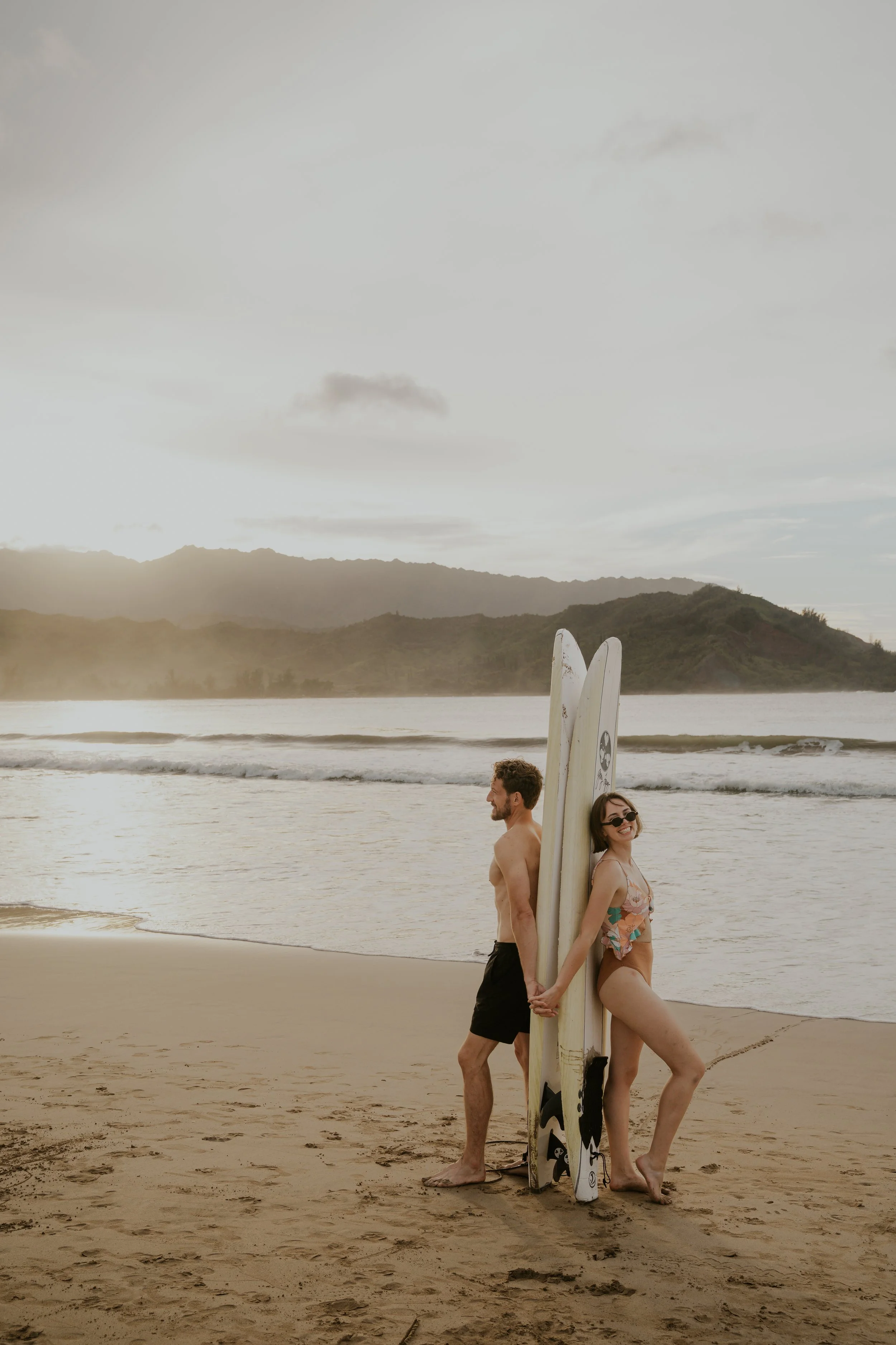 A couple standing on the beach back-to-back holding hands, with surfboards leaning against the woman, during sunset or sunrise with hills in the background.