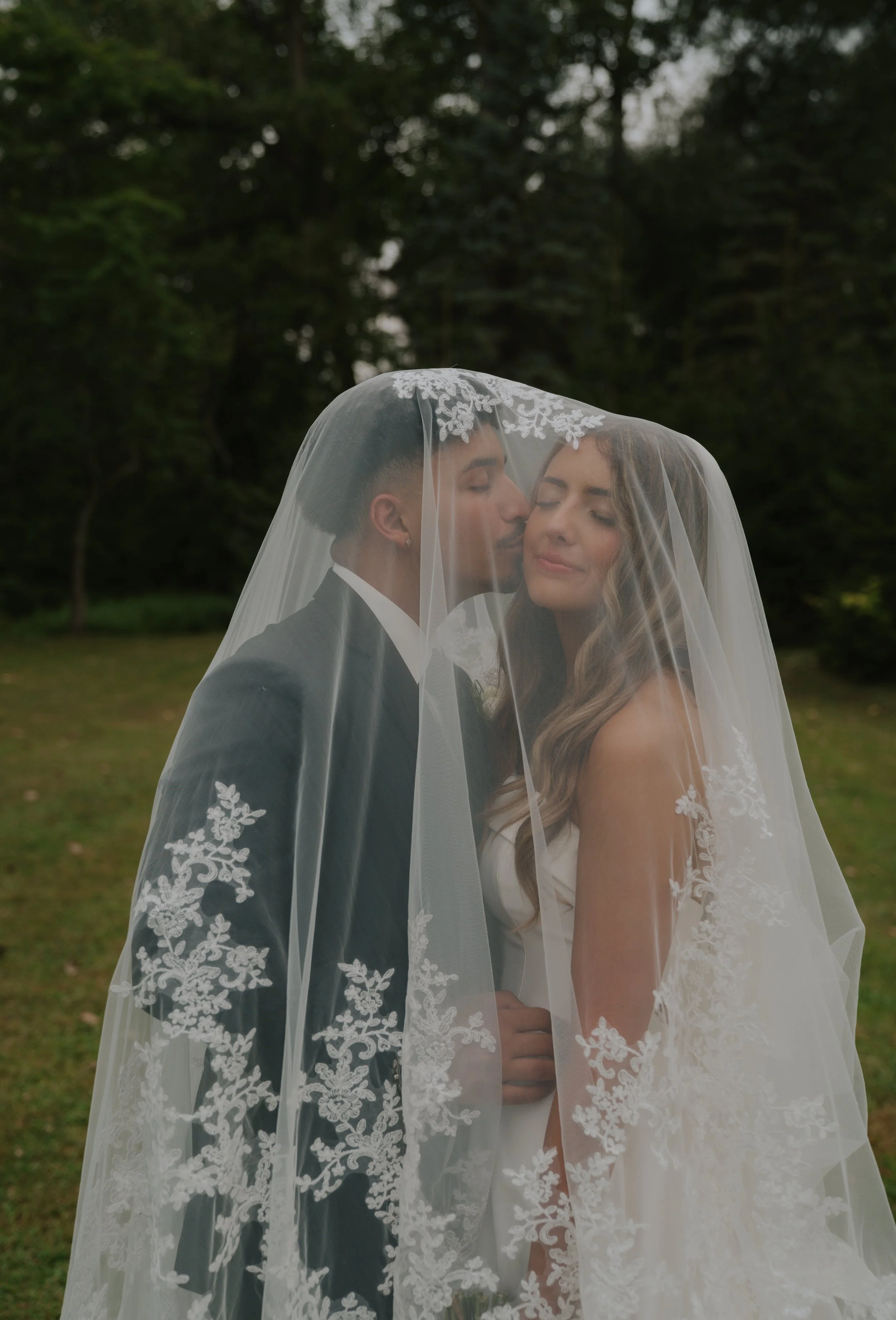 A bride and groom are under a veil, with the groom kissing the bride's forehead, outdoors on a grassy area with trees in the background.