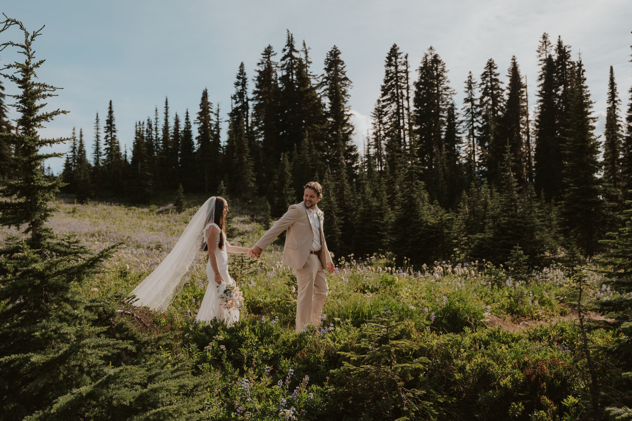 A bride and groom strolling hand-in-hand through a lush forest clearing with tall evergreen trees and wildflowers, bright sky overhead.