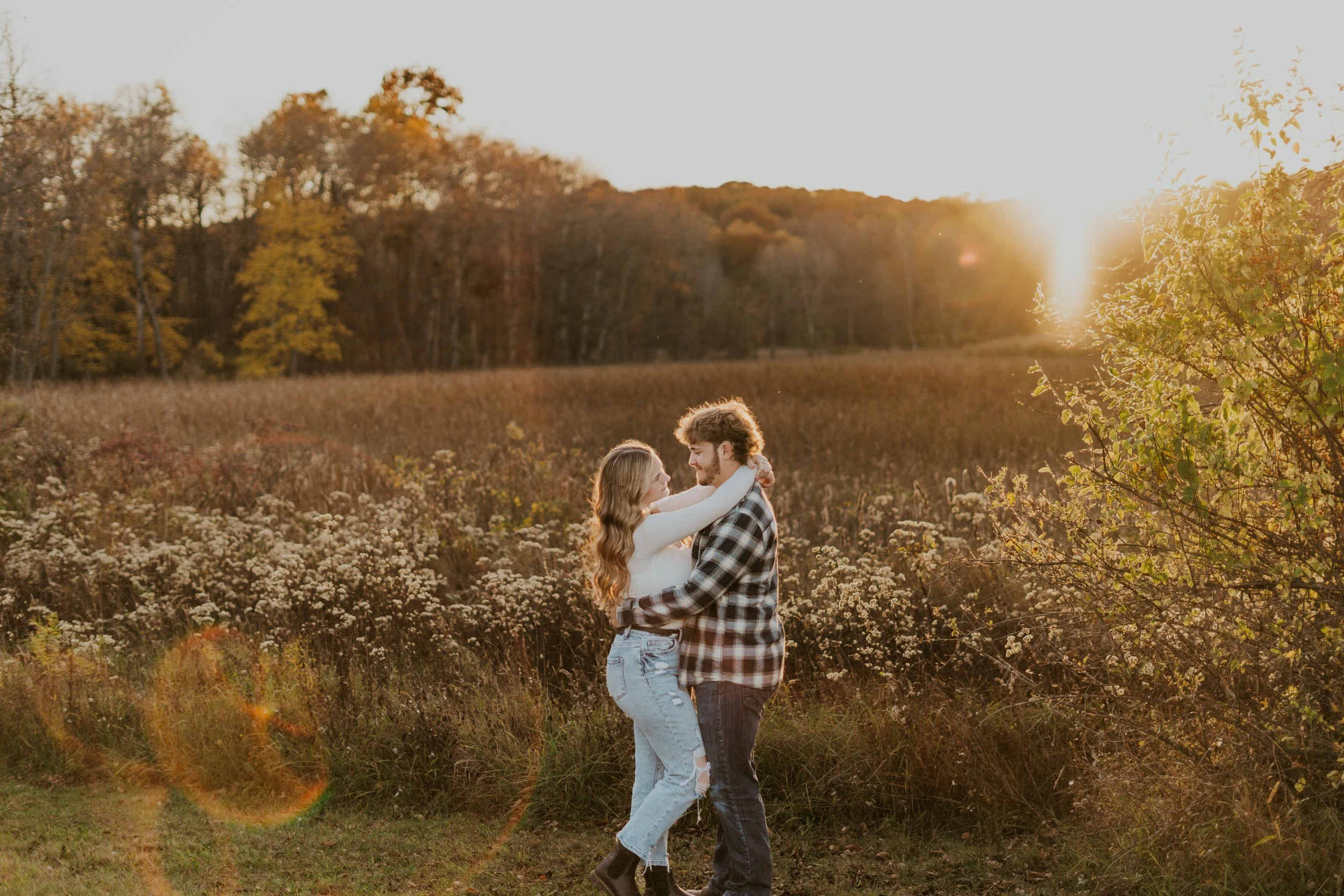 A couple embracing in a field during sunset with trees in the background.