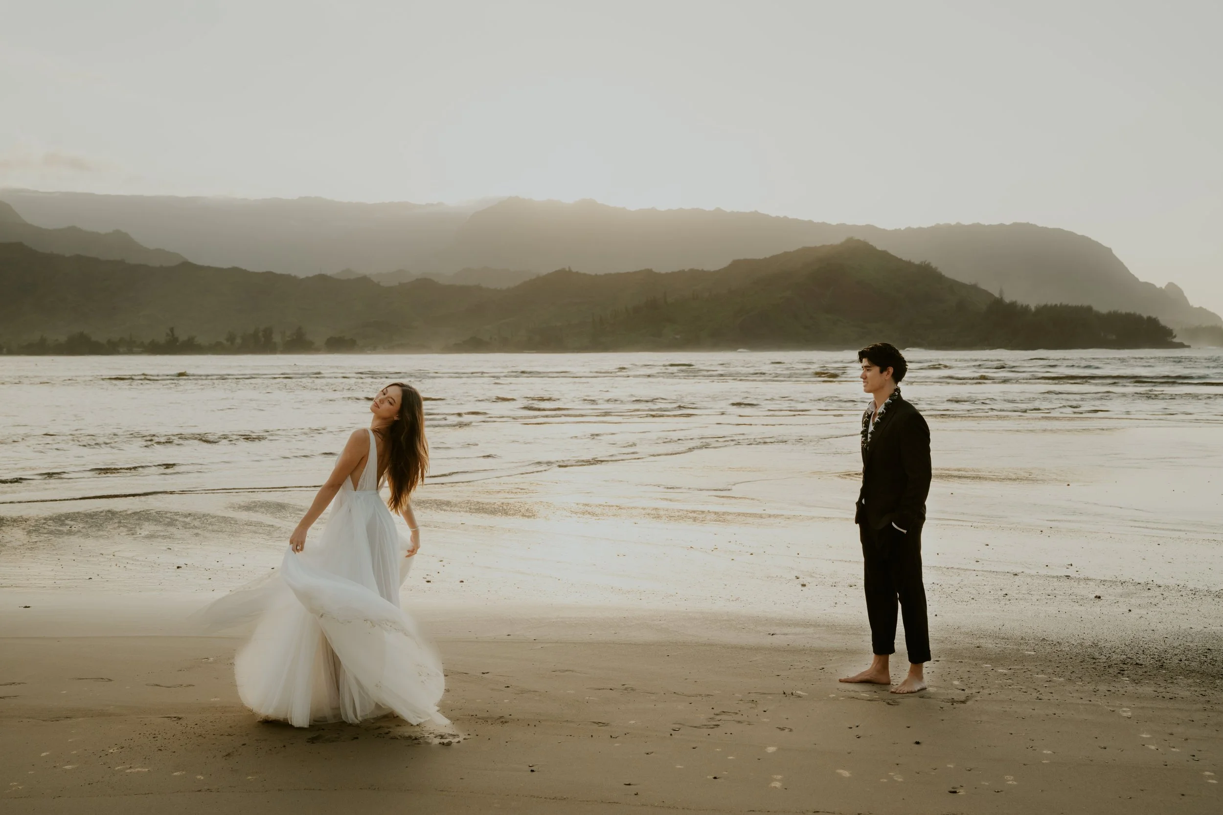 A woman in a white wedding dress and a man in a black suit stand barefoot on the beach during sunset, with mountains in the background.
