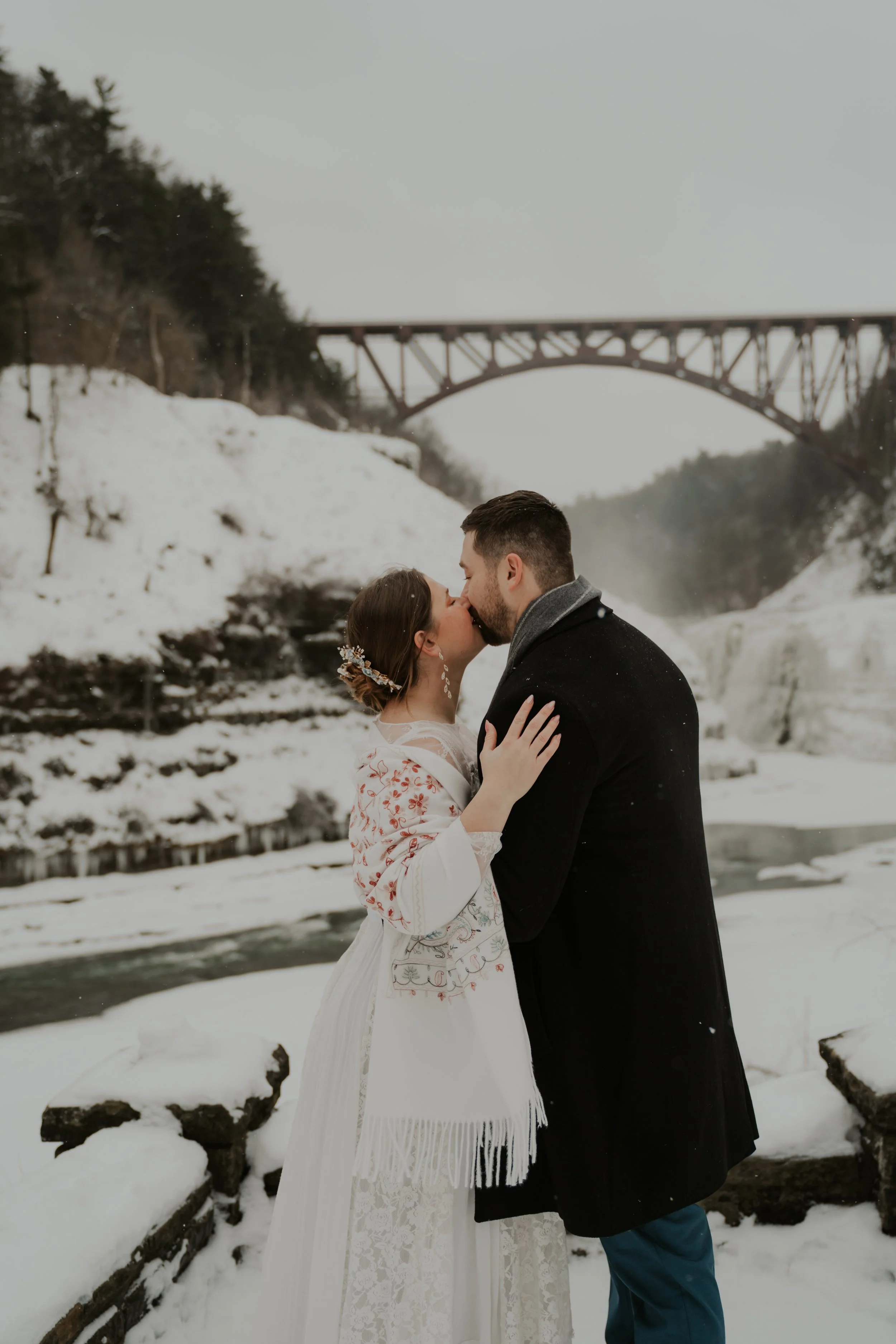 A couple sharing a kiss in a snowy landscape with a river, snow-covered rocks, trees on a hillside, and a bridge in the background.