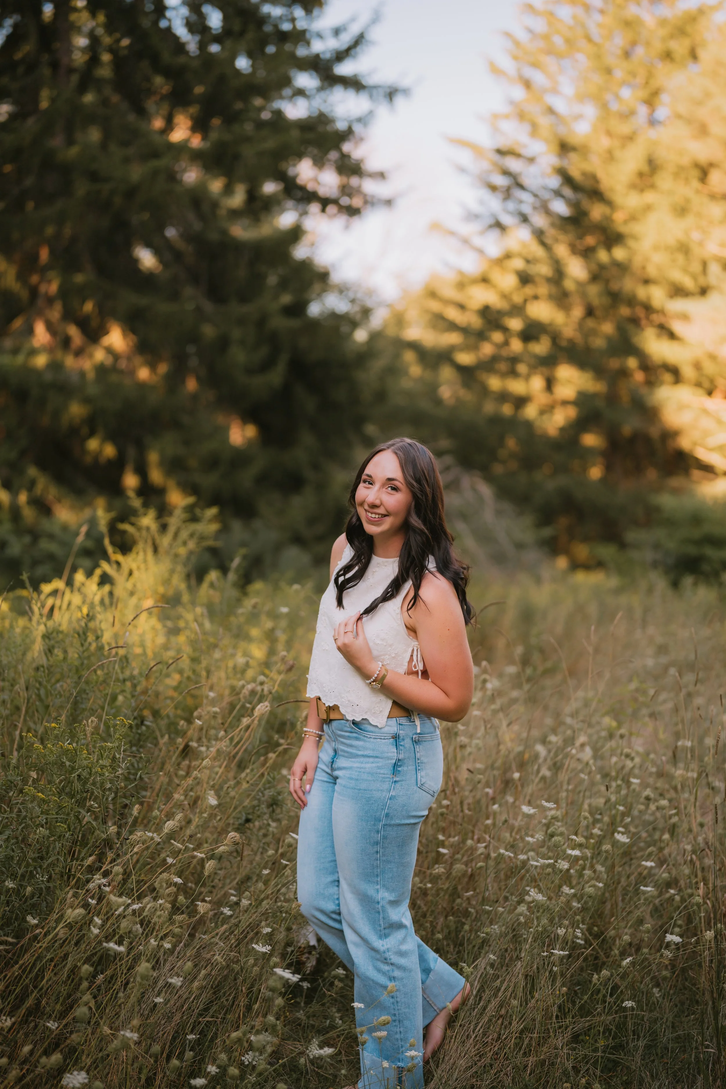A young woman with long dark hair smiling and standing in a grassy field during golden hour, surrounded by trees.