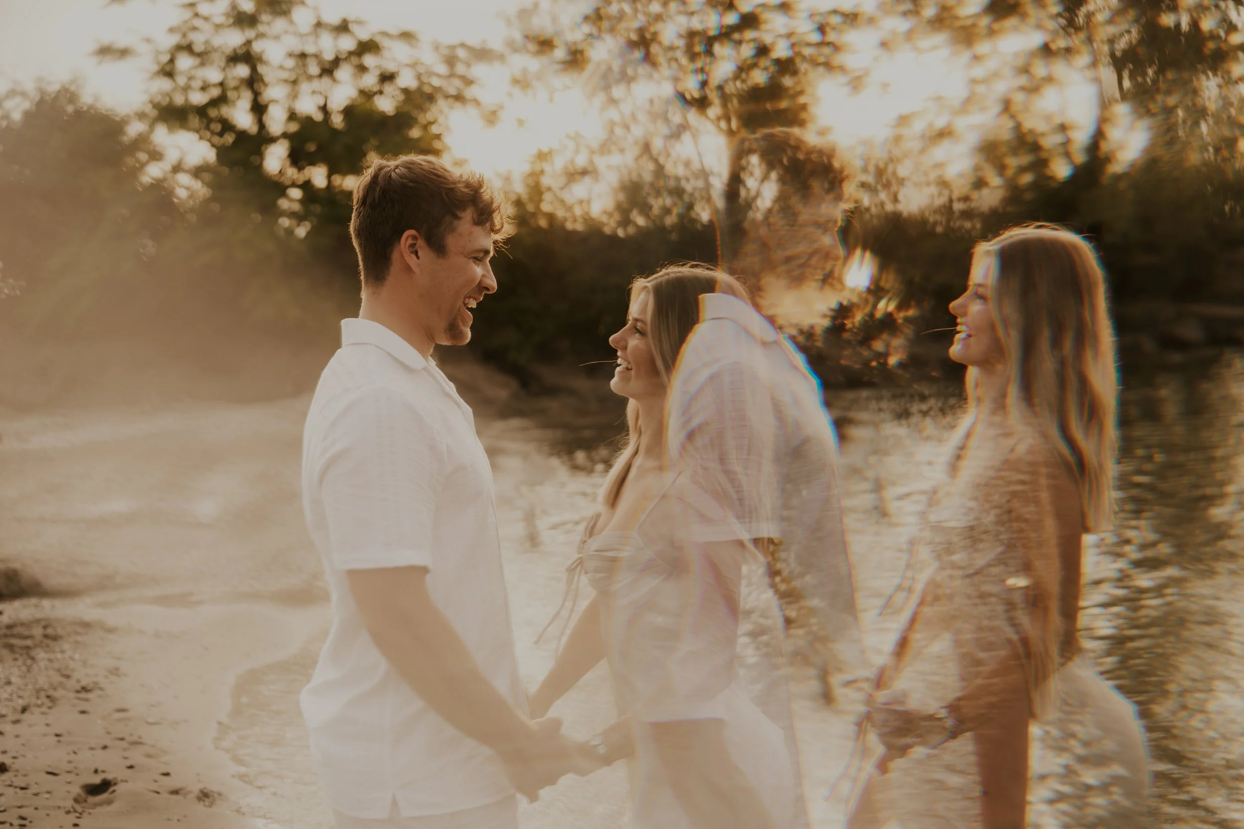A couple holding hands and smiling at each other at the beach with a double exposure of a woman in a dress and a man in a shirt, creating a layered effect during sunset.