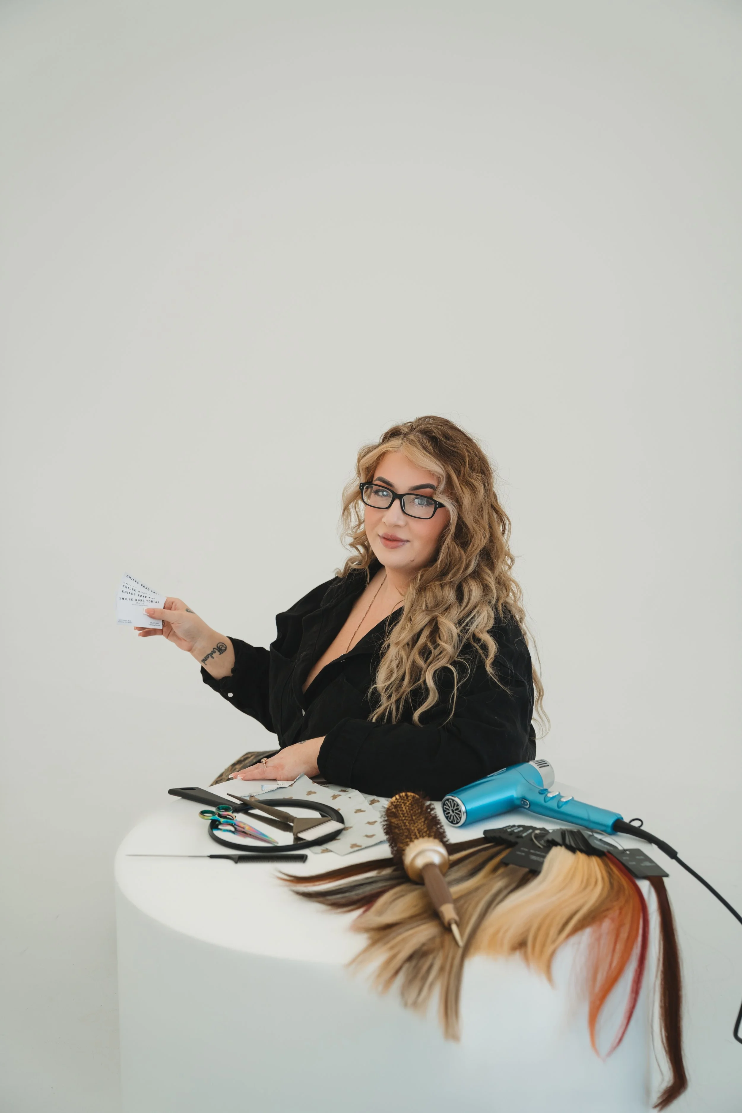 A woman with curly blonde hair, glasses, and a black shirt sitting at a white table with hair styling tools, colorful hair extensions, and a hairdryer, holding a card in her hand.