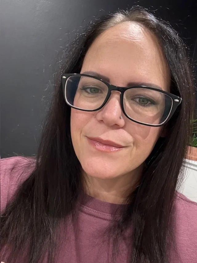 A woman with long dark hair, glasses, and a nose piercing, wearing a pink shirt, smiling softly in front of a dark background.