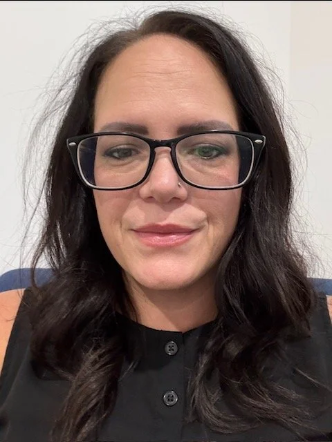 A woman with long dark hair wearing black-rimmed glasses and a black sleeveless top, smiling faintly, against a neutral background.