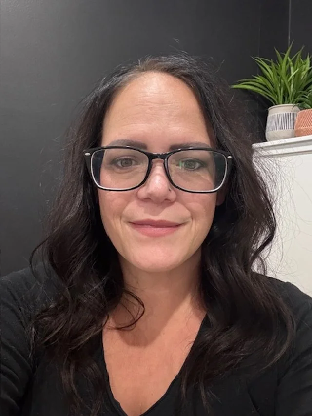 A woman with dark wavy hair wearing glasses and a black top, smiling indoors with a black wall and potted plant in the background.