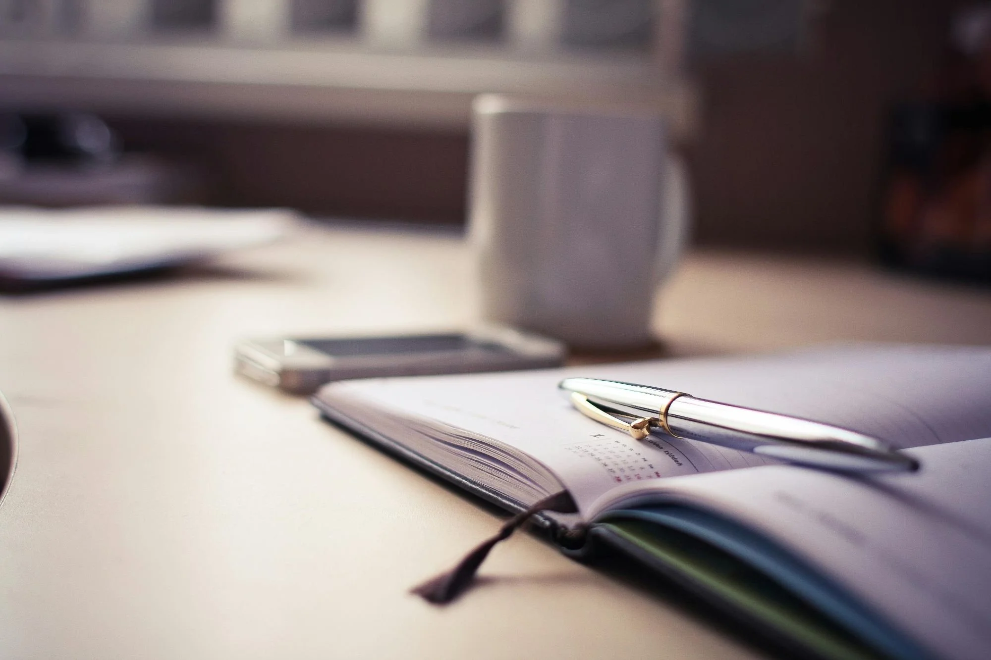 Close-up of a desk with an open planner, a pen, a smartphone, and a coffee mug in the background.
