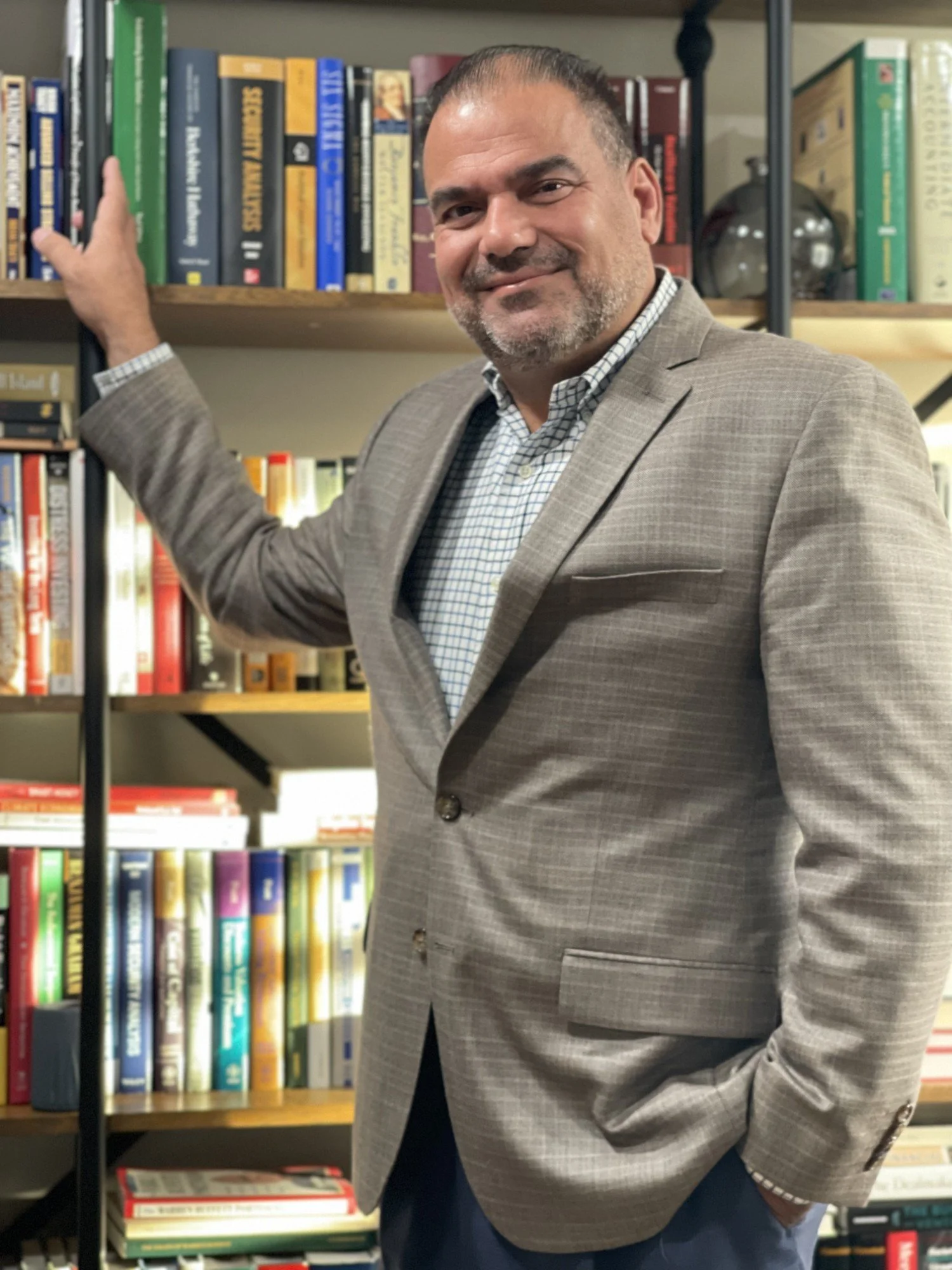 A man in a beige checked blazer and a checkered shirt stands in front of a bookshelf, smiling and resting his hand on the shelf.