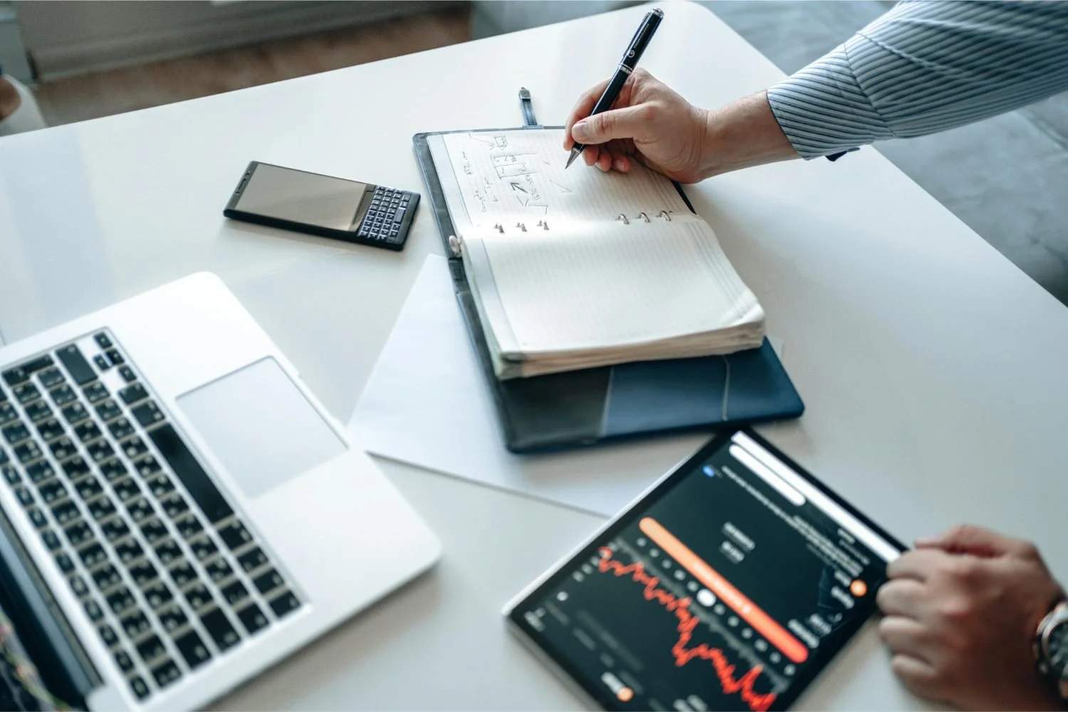 Businessperson working at a desk with a laptop, a smartphone, a notebook, and a tablet displaying a graph.