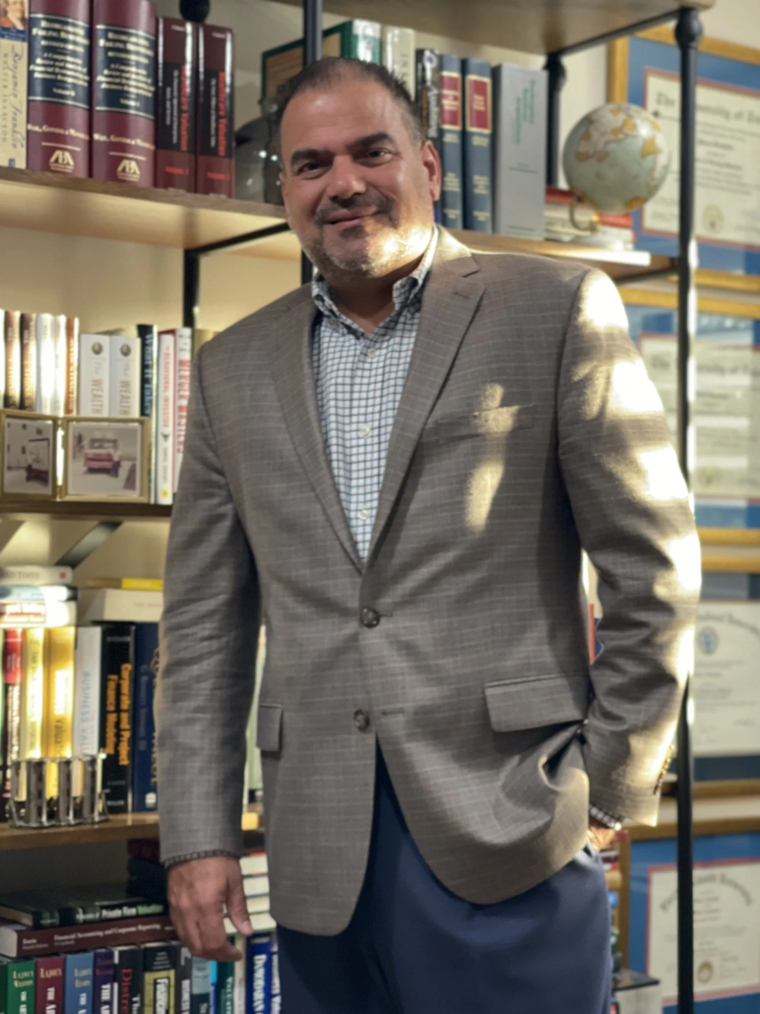 A man in a gray checked blazer and blue pants standing in front of bookshelf filled with books and a globe, smiling at the camera.