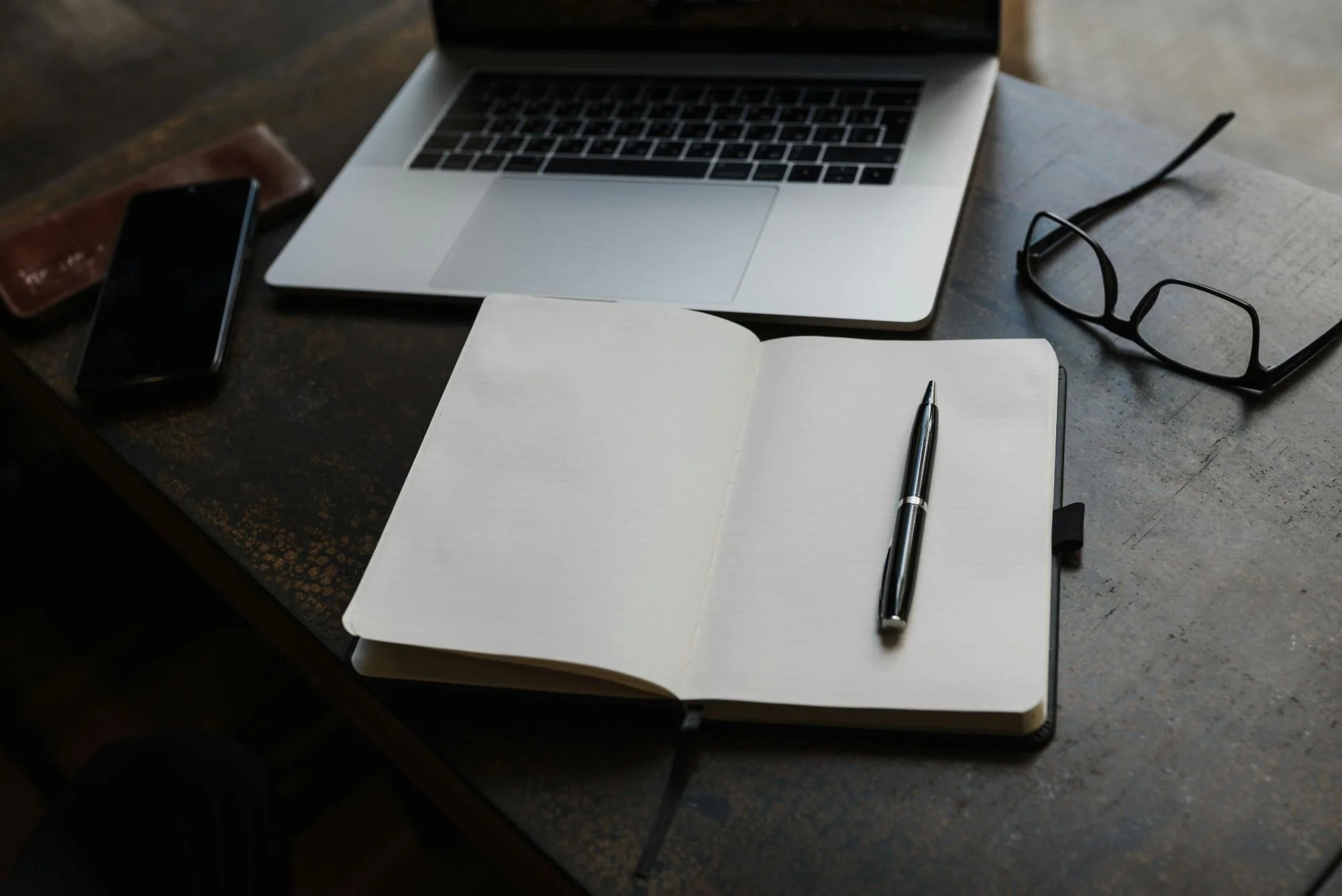 A notebook with a pen on an open page on a work desk. Surrounding the notebook are a laptop, a pair of black glasses, a smartphone, and a brown wallet.