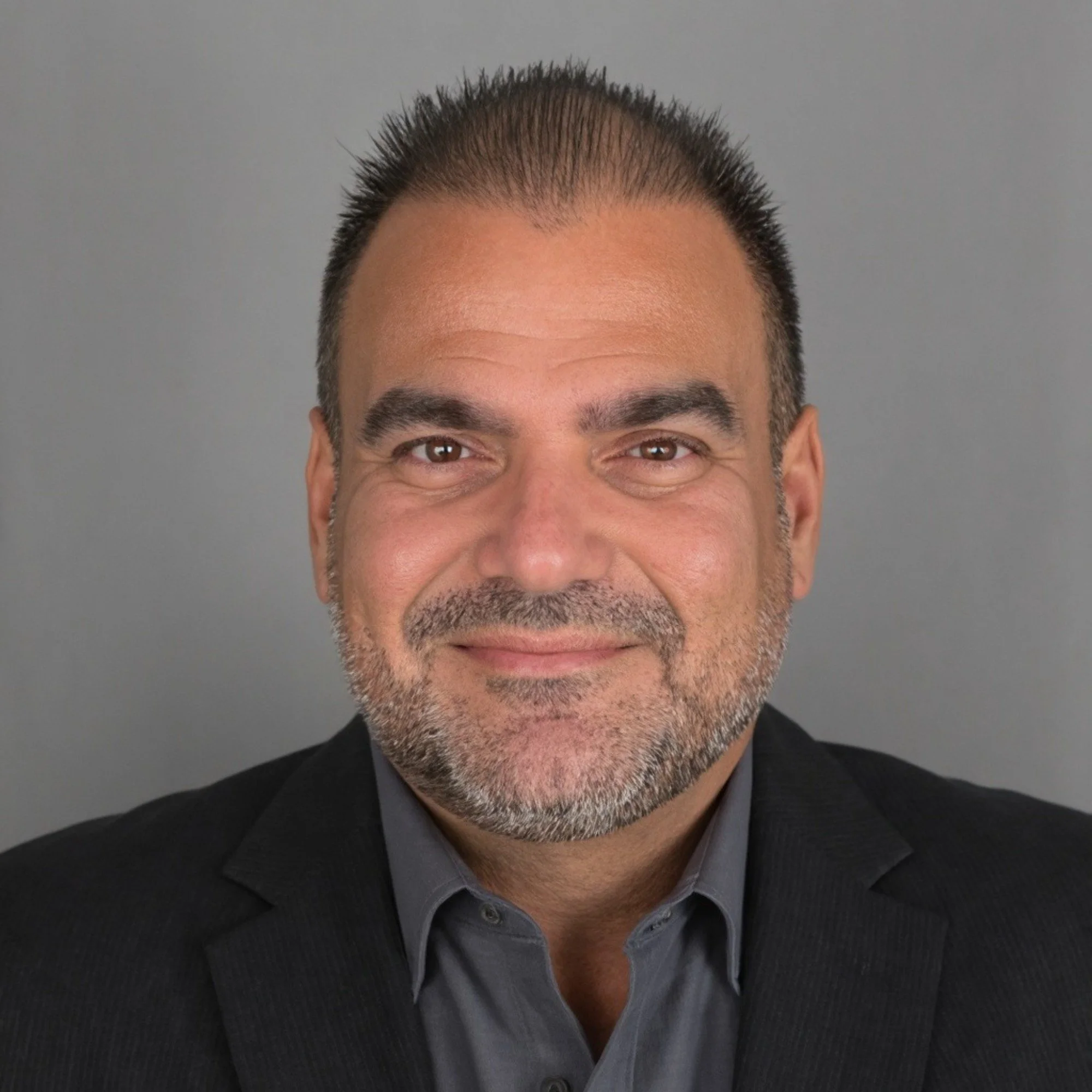 Headshot of a middle-aged man with short dark hair, gray beard, wearing a dark blazer and gray shirt against a plain gray background.