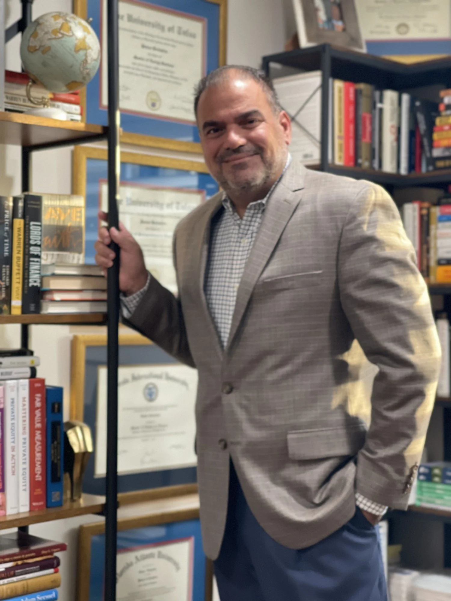 A man in a beige blazer and checkered shirt standing in a bookstore or library, holding a black cane, with books and framed certificates on the shelves behind him.