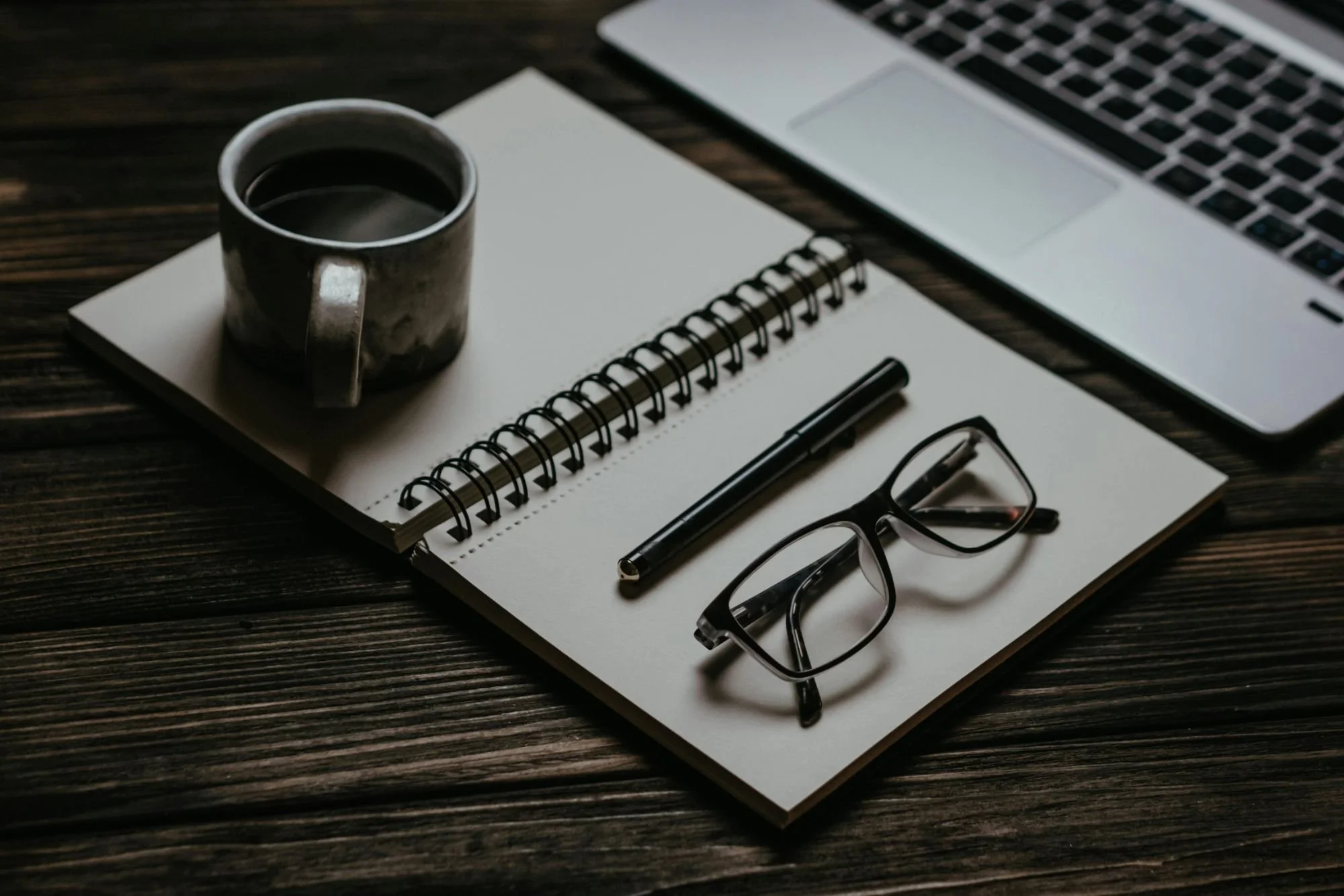 A desk scene with a cup of coffee, an open spiral notebook, a black pen, a pair of eyeglasses, and a laptop on a wooden surface.