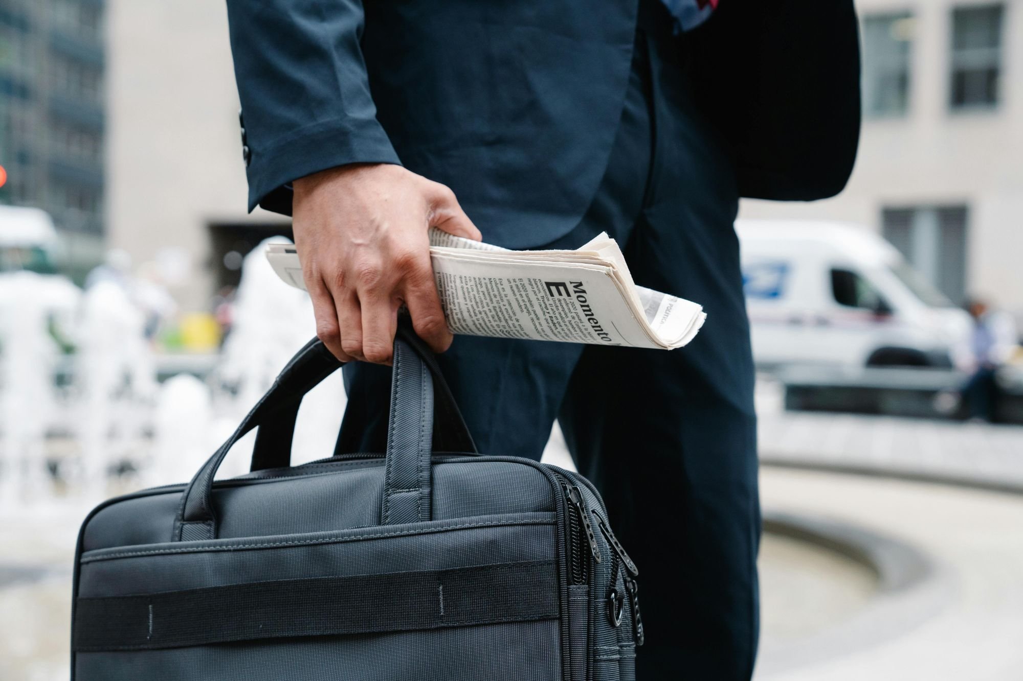 A person in a dark suit holding a folded newspaper and a black briefcase in an urban setting.