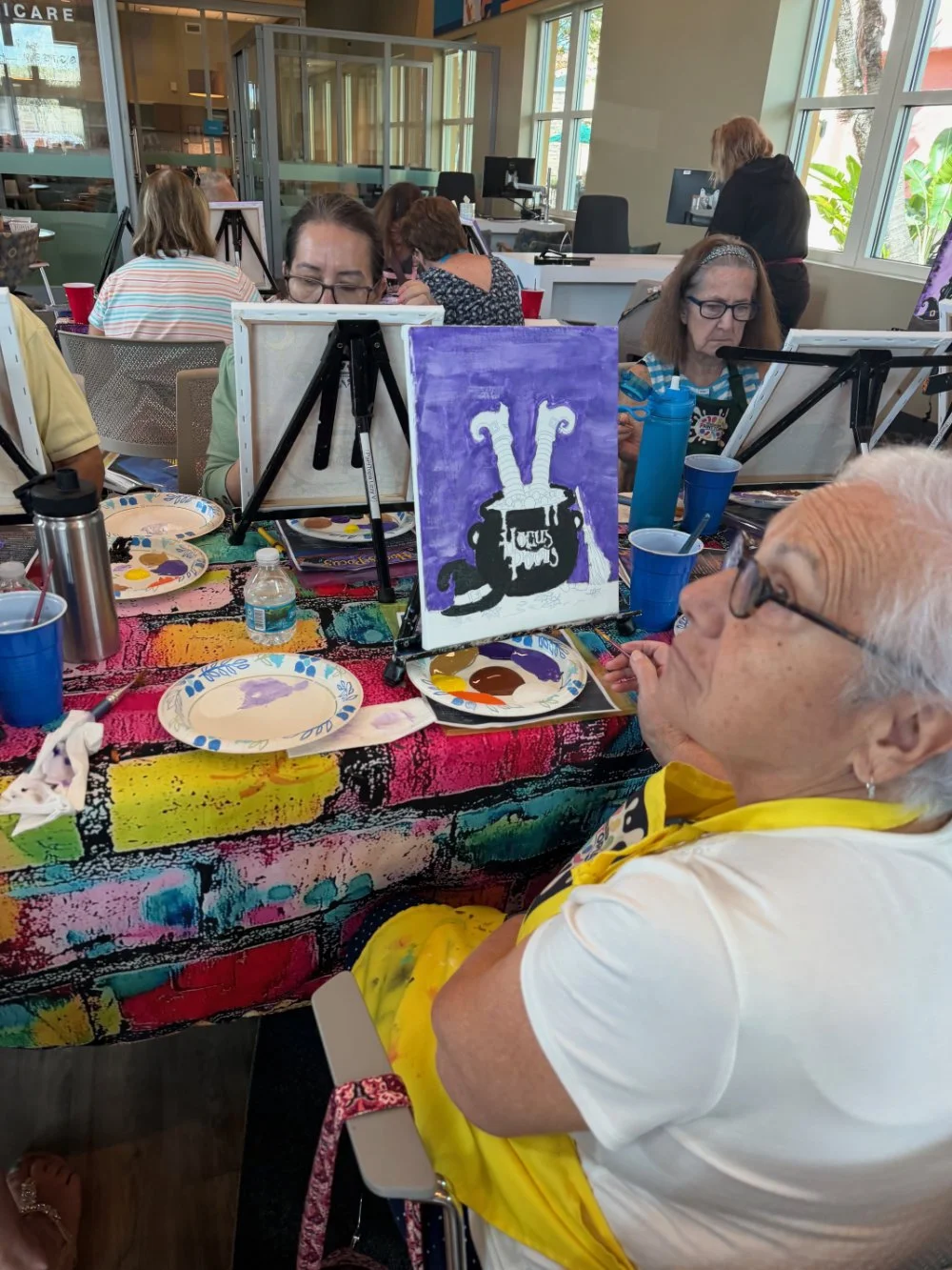 A group of people participating in a paint and sip event, with an elderly woman in the foreground wearing glasses and a yellow apron, sitting at a table covered with colorful paint splatters, plates with paint, and beverages. They are painting a purp