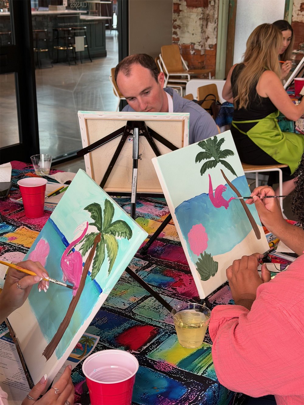 People painting tropical scene with palm trees and flamingos on canvases at a group art event.