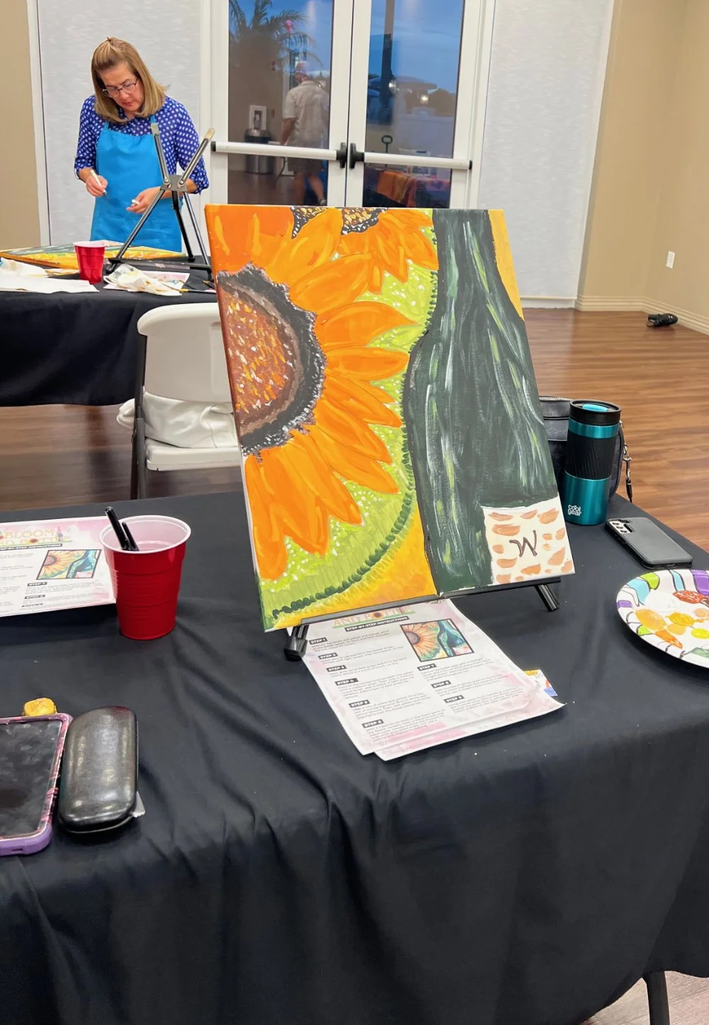 A partially finished painting of a sunflower on an easel, with bright yellow petals and a dark center, displayed on a table covered with black cloth. In the background, a woman wearing a blue apron and polka-dot blouse is working on her own art proje