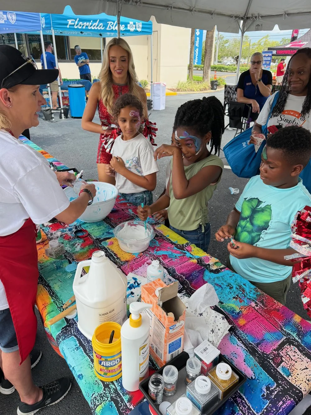 Children at an outdoor face painting booth with a colorful tablecloth, various craft supplies, and a woman demonstrating face painting, with onlookers and a blue Florida Blue tent in the background.
