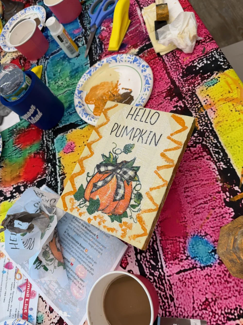 Festive table setup with a colorful tablecloth, decorated with a 'Hello Pumpkin' sign featuring a pumpkin illustration, paint supplies, paper cups with drinks, and various craft materials.