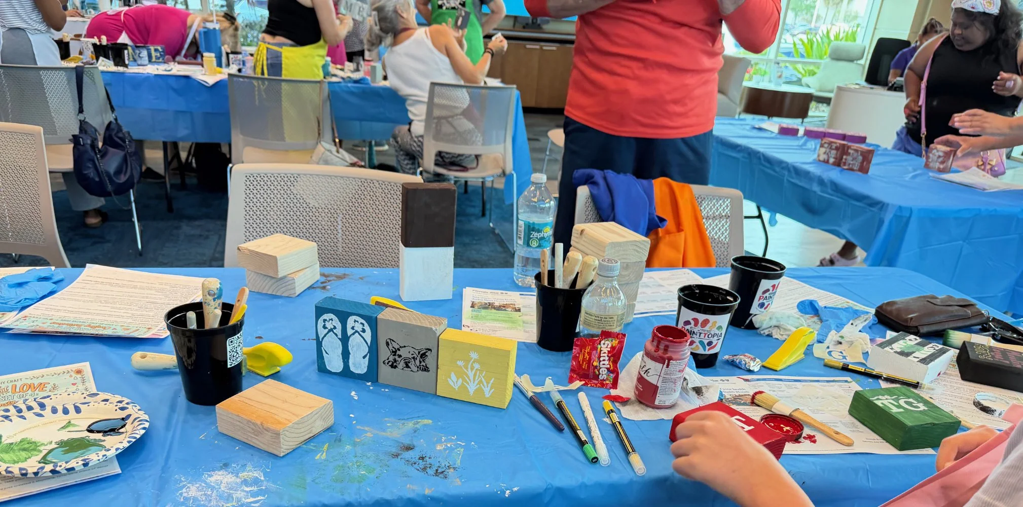 A colorful indoor crafting event with tables covered in blue tablecloths, various art supplies, and people engaged in creative activities. Some children and adults are working on projects, and there are various art materials scattered across the tabl