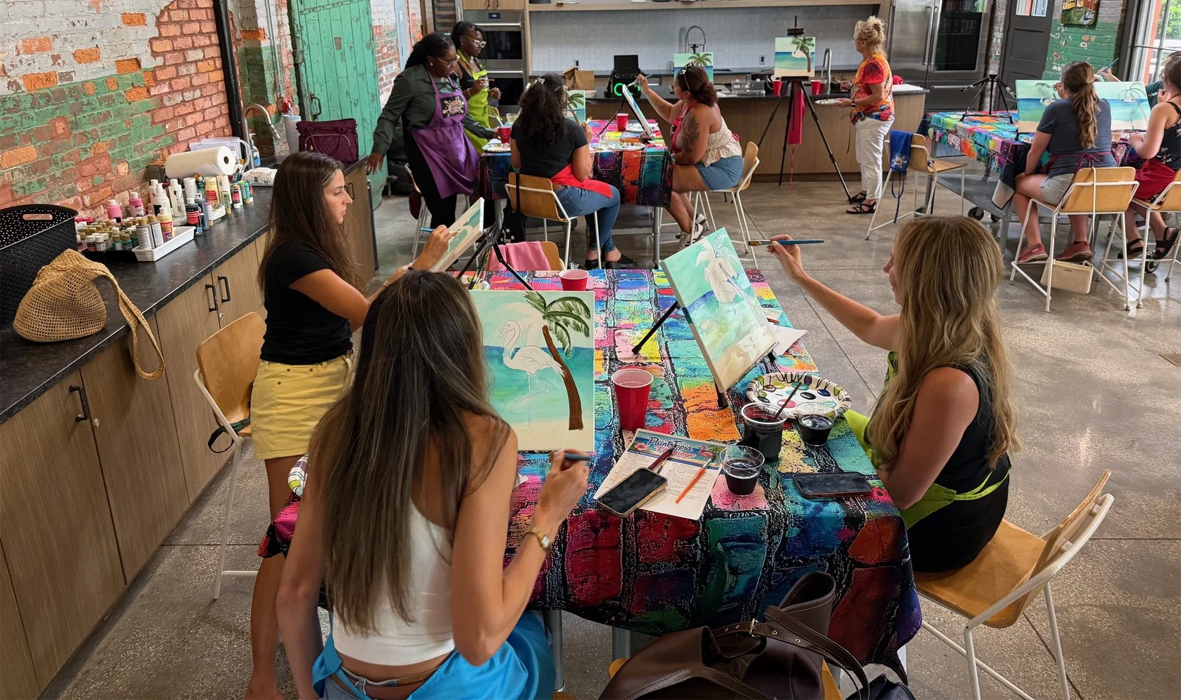 Women and girls participating in a painting class in an art studio, with some painting beach scenes featuring a swan and a palm tree.