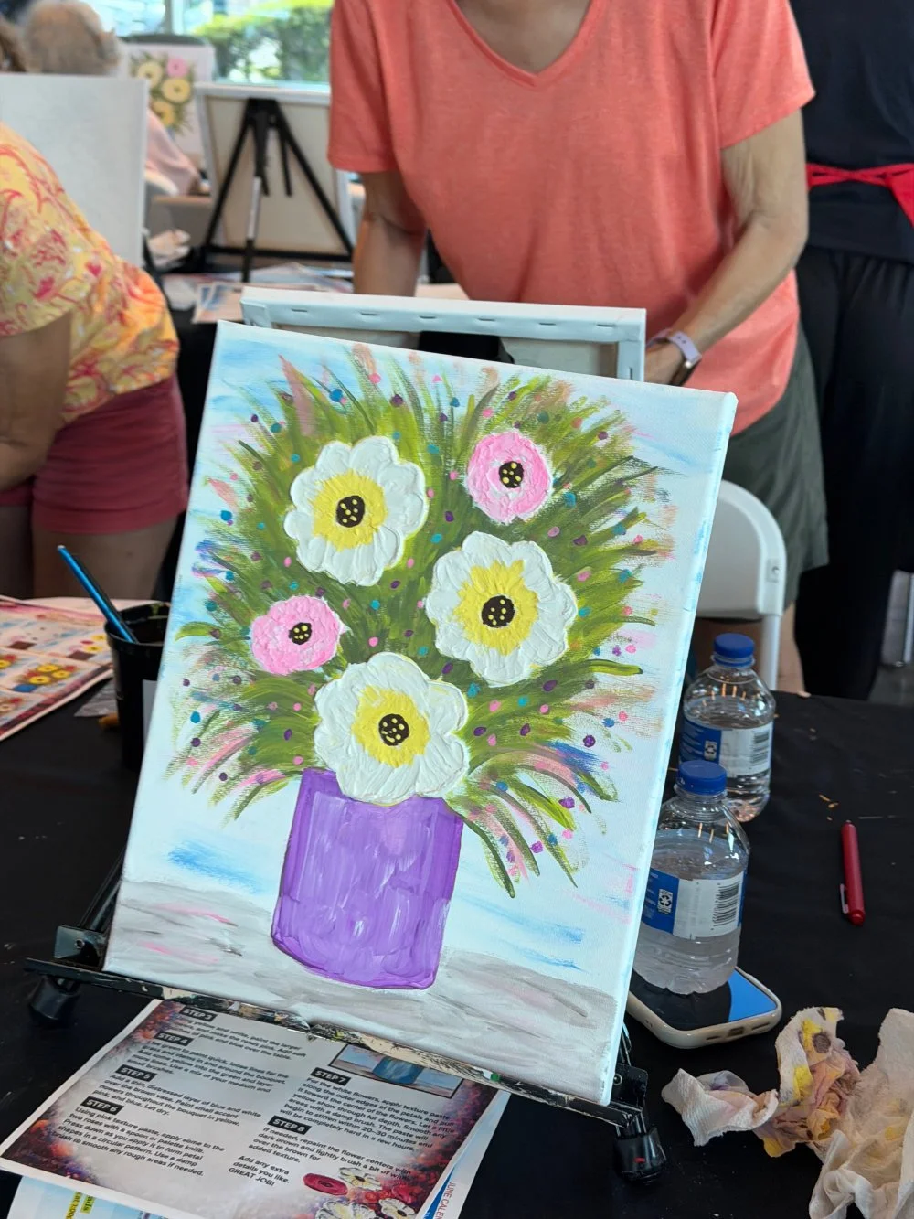 Painting of a flower arrangement in a purple vase, featuring large white and pink flowers with yellow and black centers, on an easel amid painting supplies.