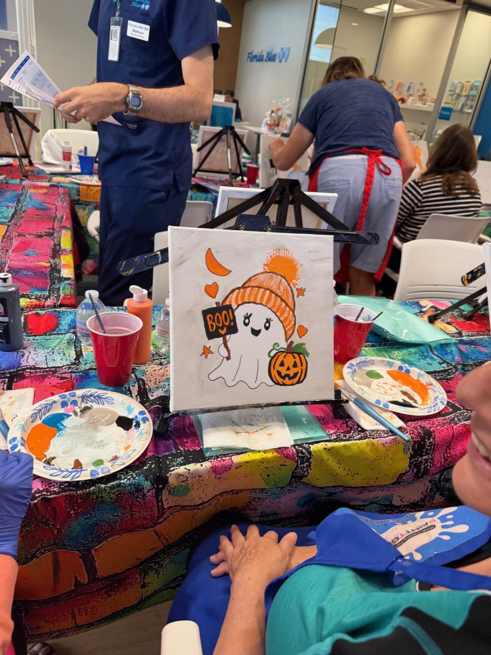 A Halloween-themed painting of a cute ghost wearing a knit hat, holding a sign that says "BOO!", with a pumpkin and various Halloween decorations around it, on an easel at a colorful arts and crafts table.
