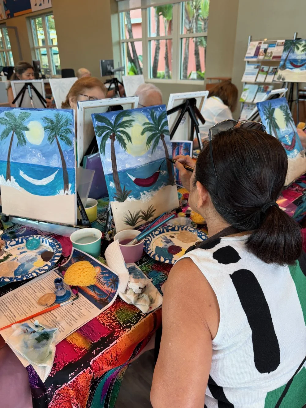 People participating in an indoor painting class, creating tropical beach scenes with palm trees, a setting sun, and ocean waves on their canvases.