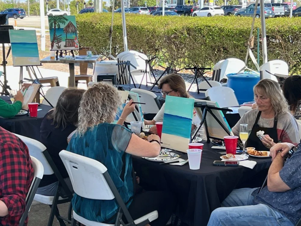 A group of people painting landscape artworks at outdoor paint and sip event. Several completed paintings of beaches and ocean scenes are visible.