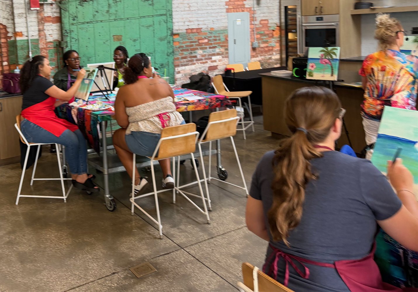 Group of women painting at a social art class in a rustic studio with brick walls and a colorful tablecloth.