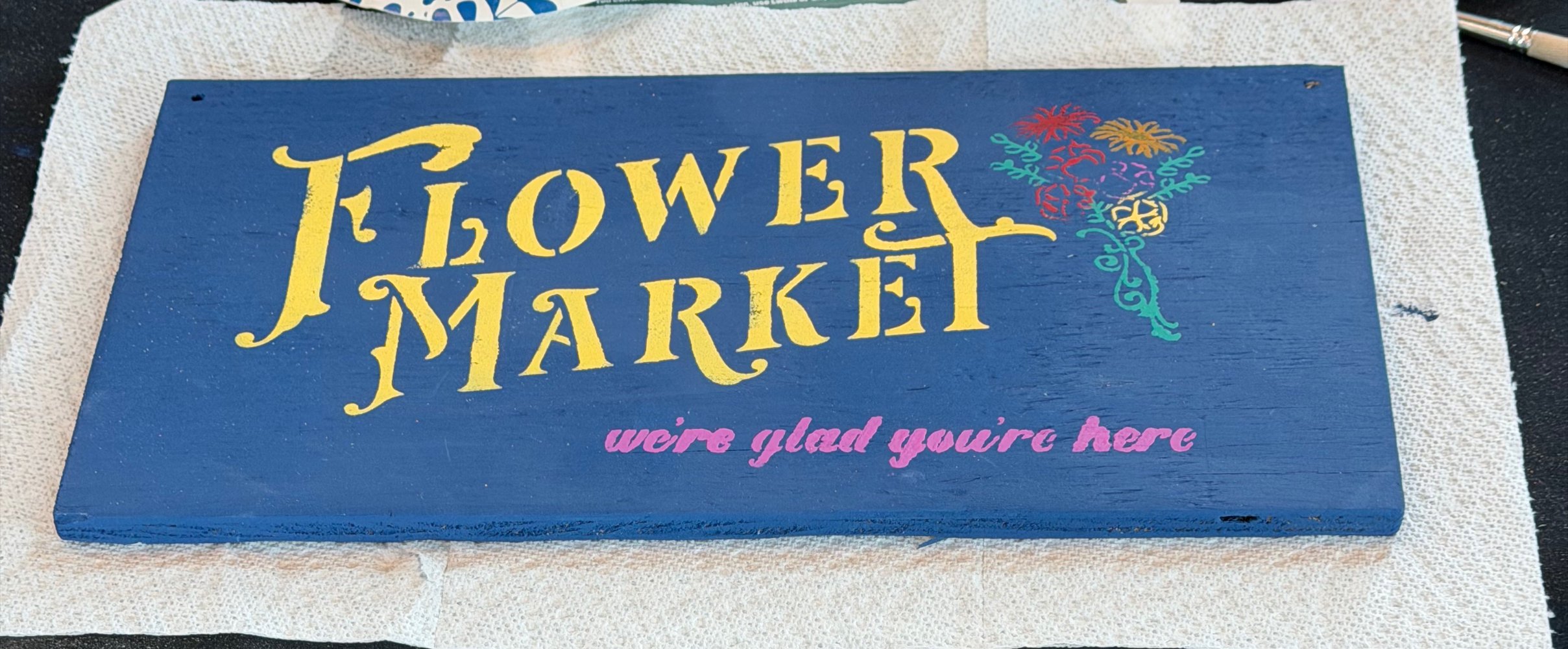 A painted wooden sign that reads 'Flower Market' with colorful flowers and the phrase 'we're glad you're here' underneath.