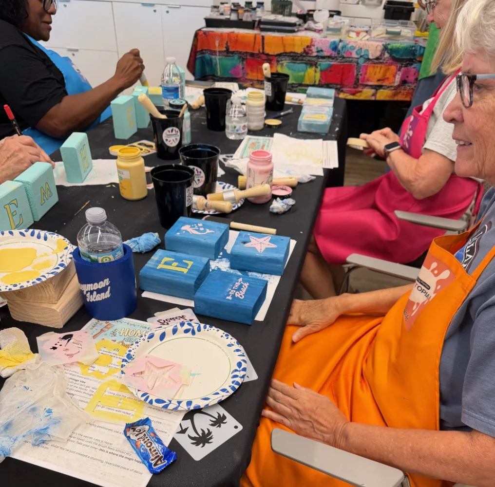 People painting wooden blocks and small canvases at a table, with paint, brushes, and decorations. There is colorful artwork and supplies in the background.