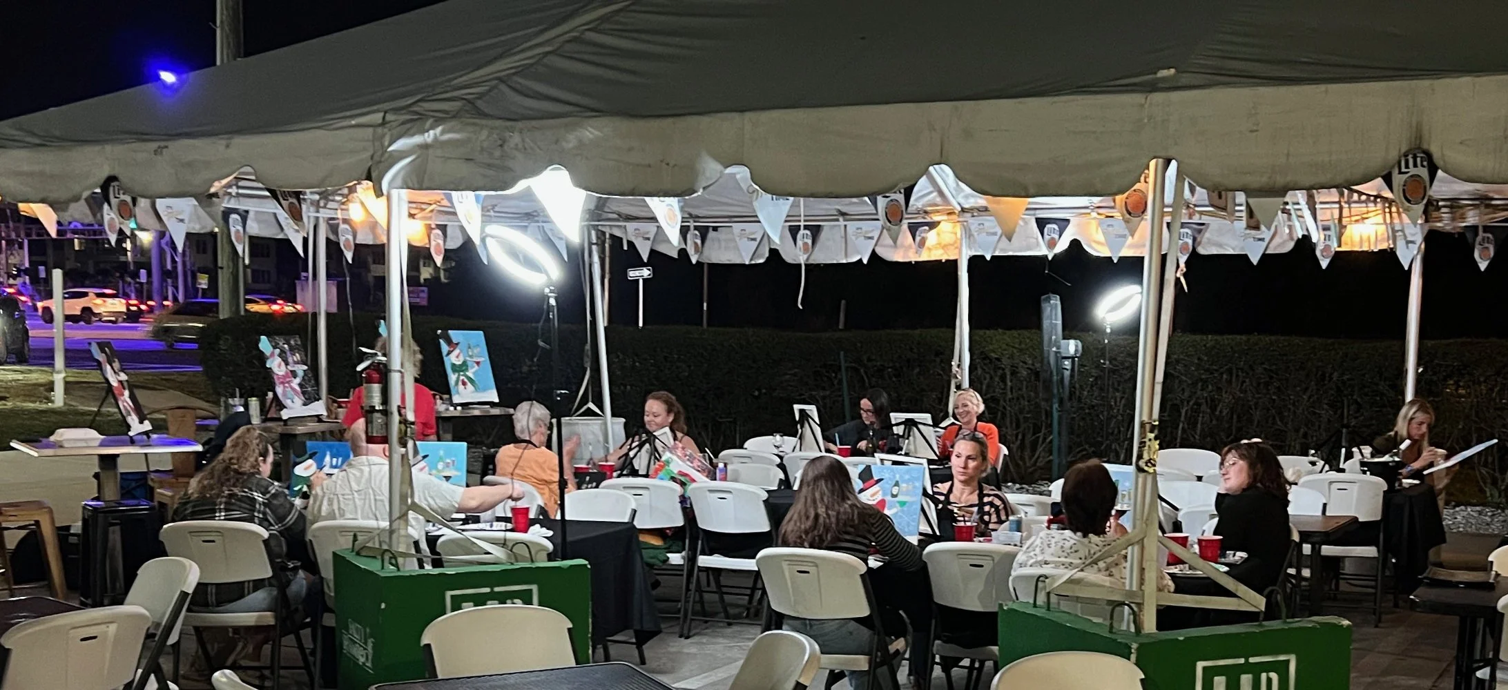Nighttime scene of an outdoor art event under a tent with people sitting at tables, some working on art projects or talking, with paintings on display and festive decorations.