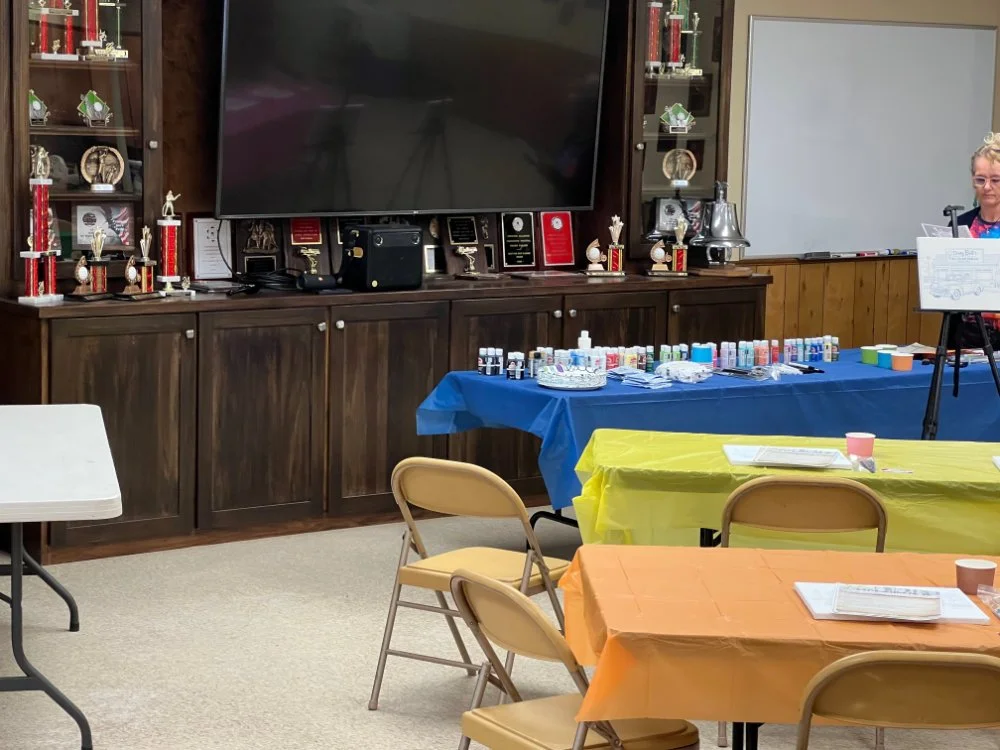 A room with tables covered in yellow, blue, and orange tablecloths, set up for a celebration or event. There are chairs, a large TV on a wooden cabinet displaying trophies and awards, and a woman standing near a music stand. The tables have cups, pap