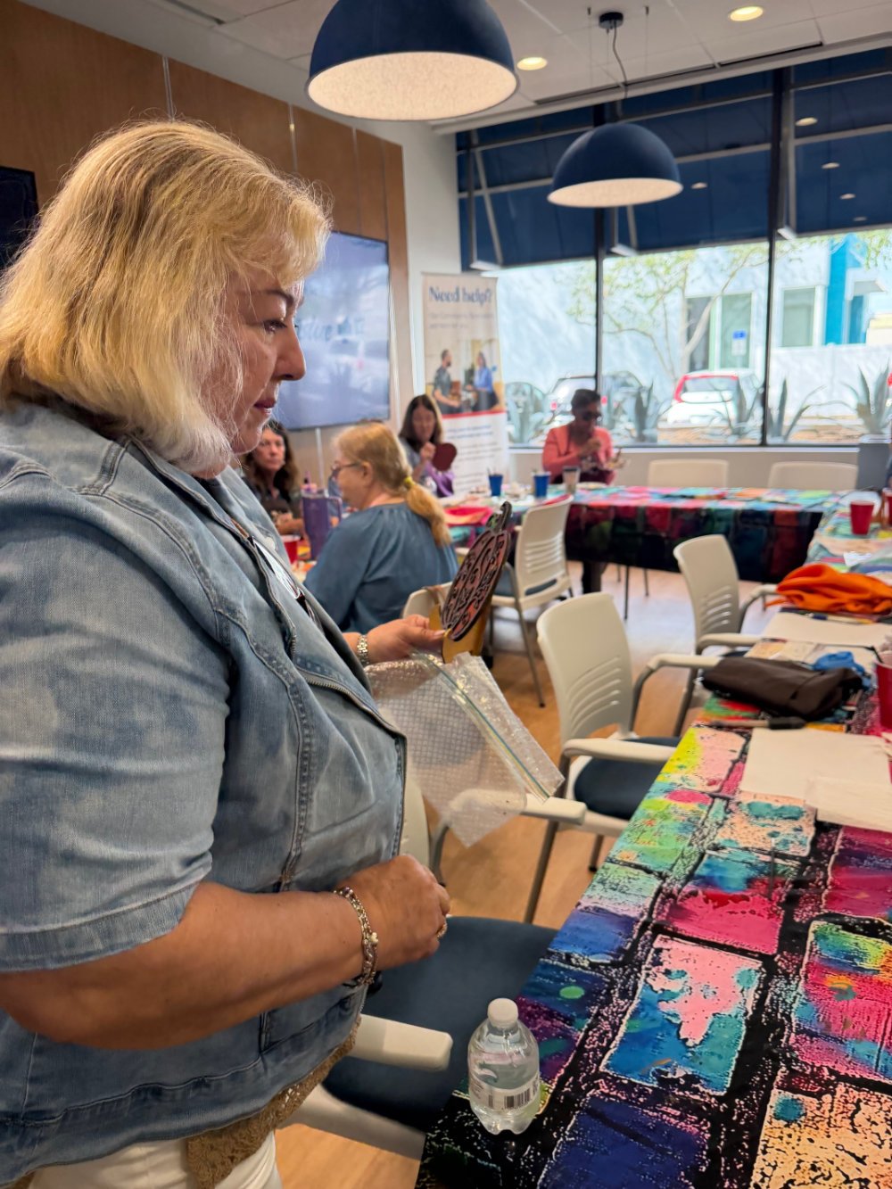A woman with blonde hair in a denim jacket is holding a painted tile and a paintbrush at a table covered with a colorful cloth in a room with other people, large windows, and blue pendant lights.
