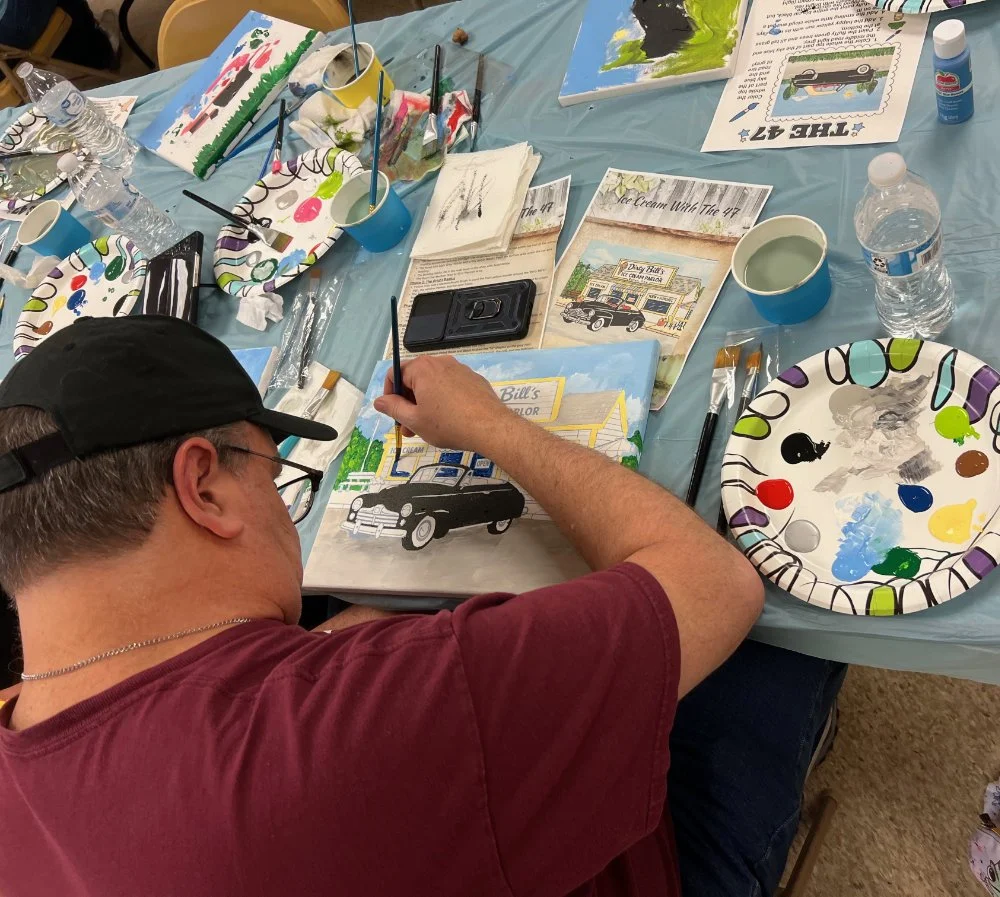 A man is painting a picture of a vintage black and white car outside a building labeled 'Bill's' with a light blue sky and green trees. The table has paint supplies, paper plates with paint, brushes, water bottles, and a camera.