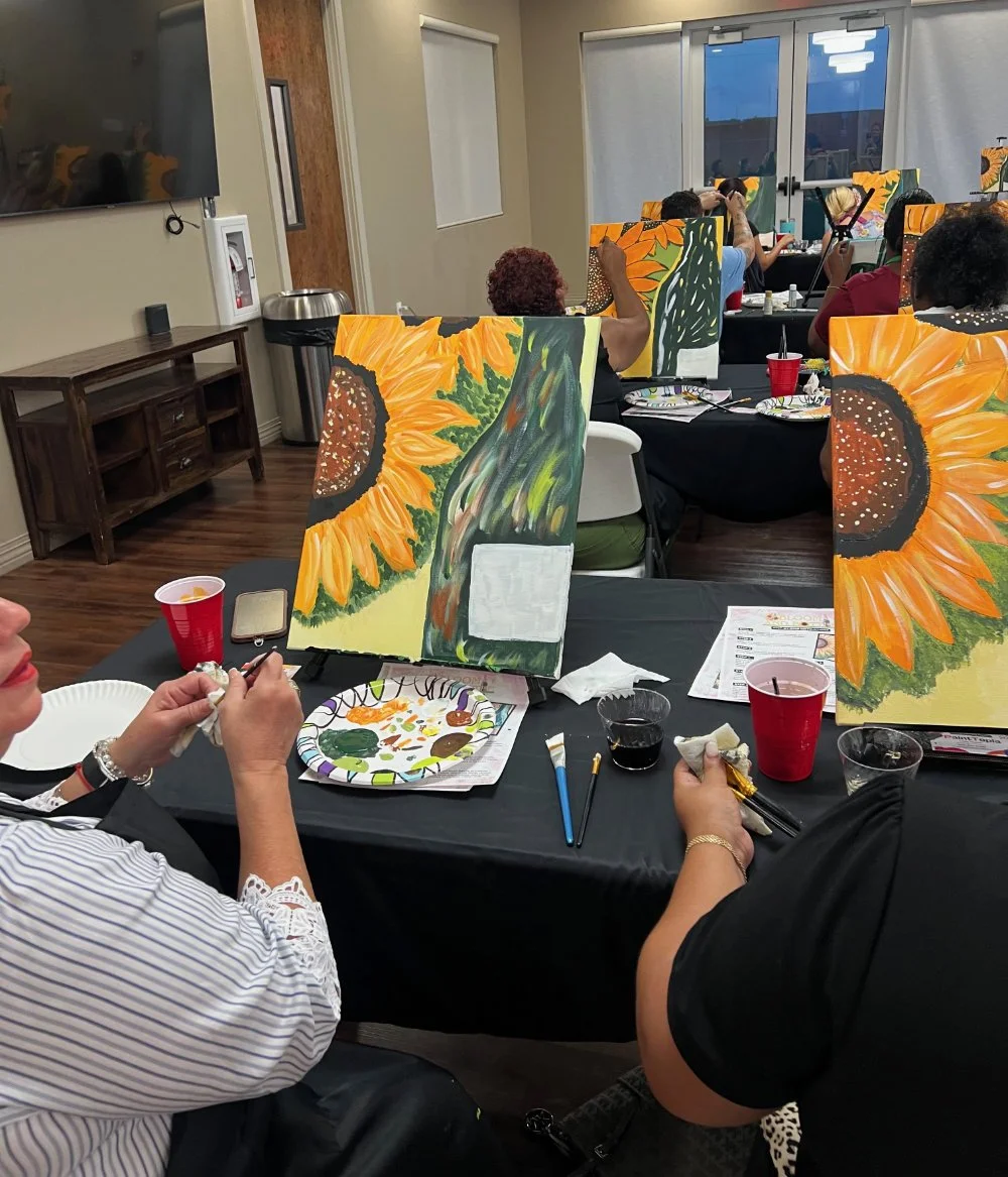 People painting sunflower artwork at an indoor art class, with paint supplies and drinks on the table.