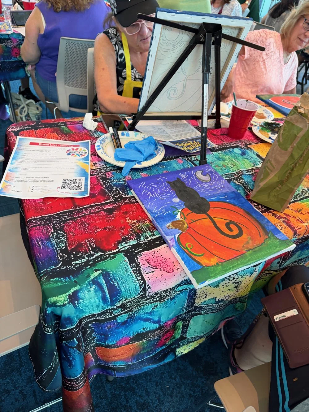 A table with a colorful abstract tablecloth displays a painting of a black cat sitting on an orange pumpkin with green vines, under a background of swirling blue sky with a crescent moon. The table is set up for a painting activity with a painting ea