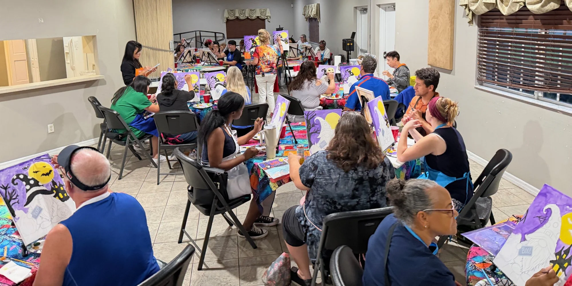 Group of people participating in a Halloween-themed painting class, seated around tables with canvases depicting spooky scenes, some with bats, a haunted house, and a moon, in a room decorated with Halloween motifs.